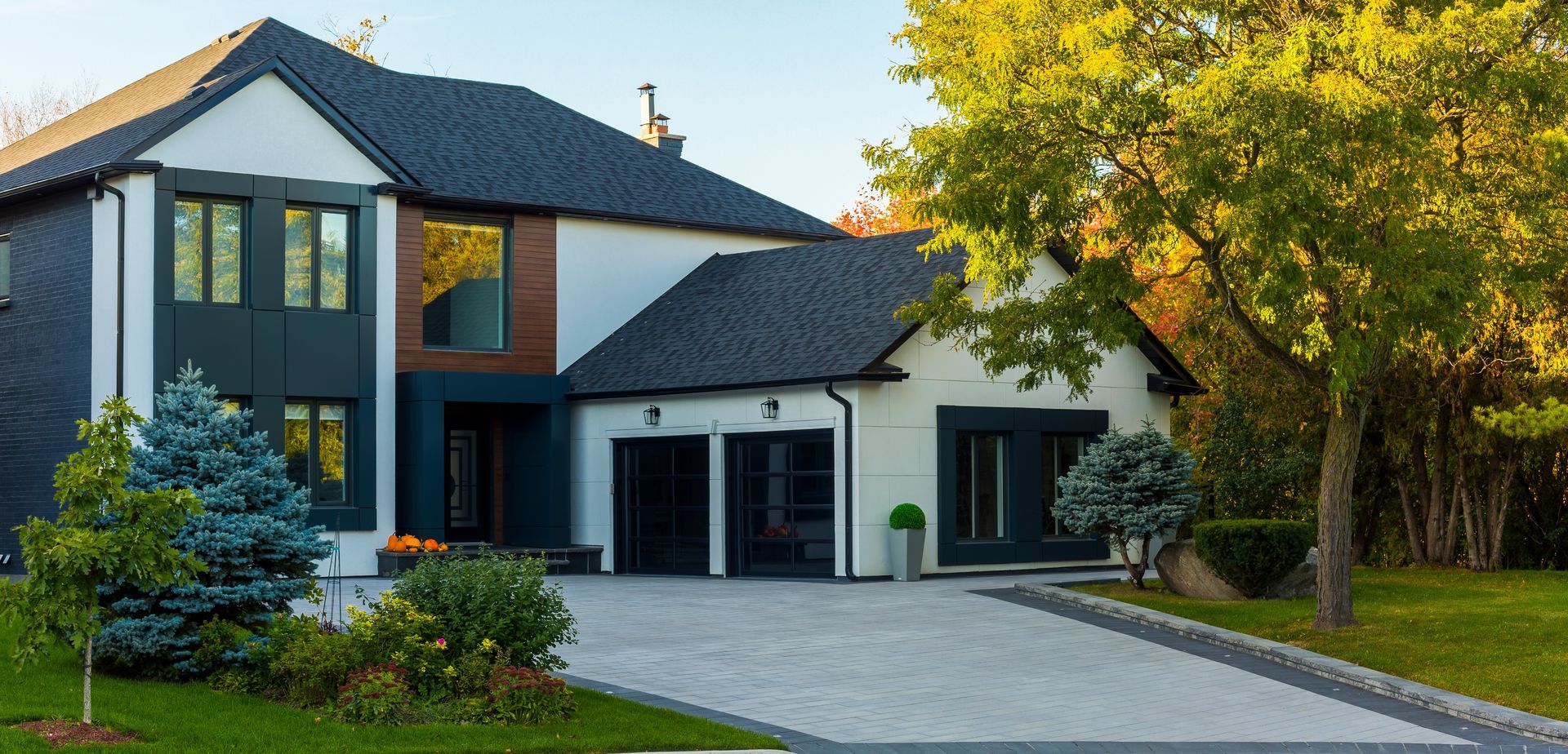 Maison moderne avec toit gris foncé, murs blancs et allée pavée. Arbres et aménagement paysager à l'avant.