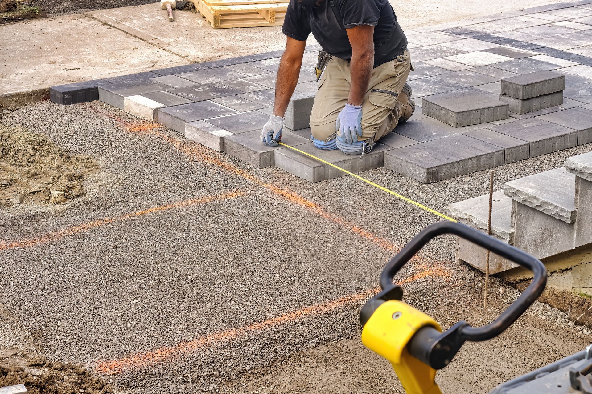 Un homme agenouillé mesure des pavés posés sur un lit de gravier. Mètre ruban, lignes orange, matériaux de construction.