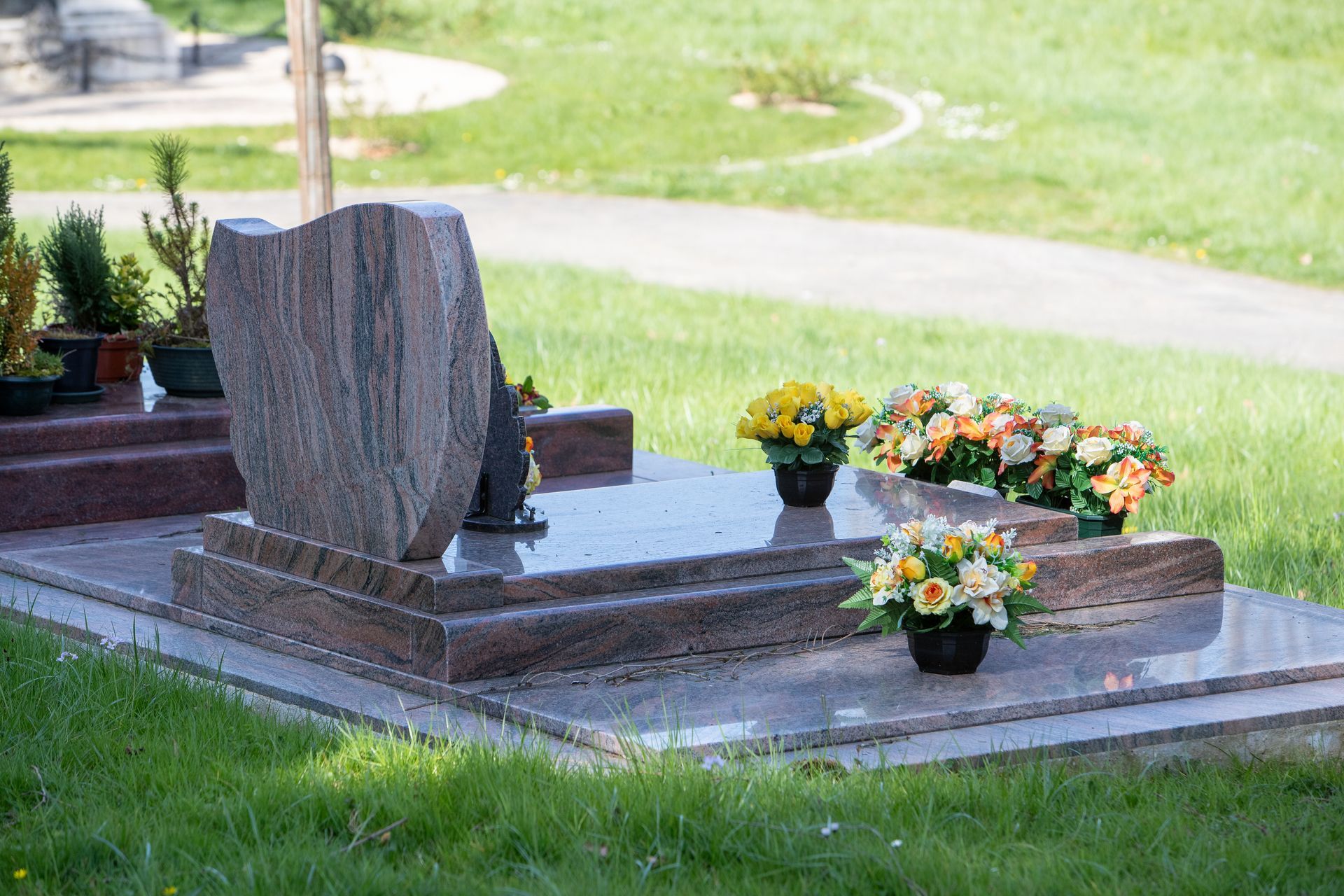 Grave with a pinkish granite headstone and base; flowers in pots sit around it, set in a grassy cemetery. Grave with a pinkish granite headstone and base; flowers in pots sit around it, set in a grassy cemetery.