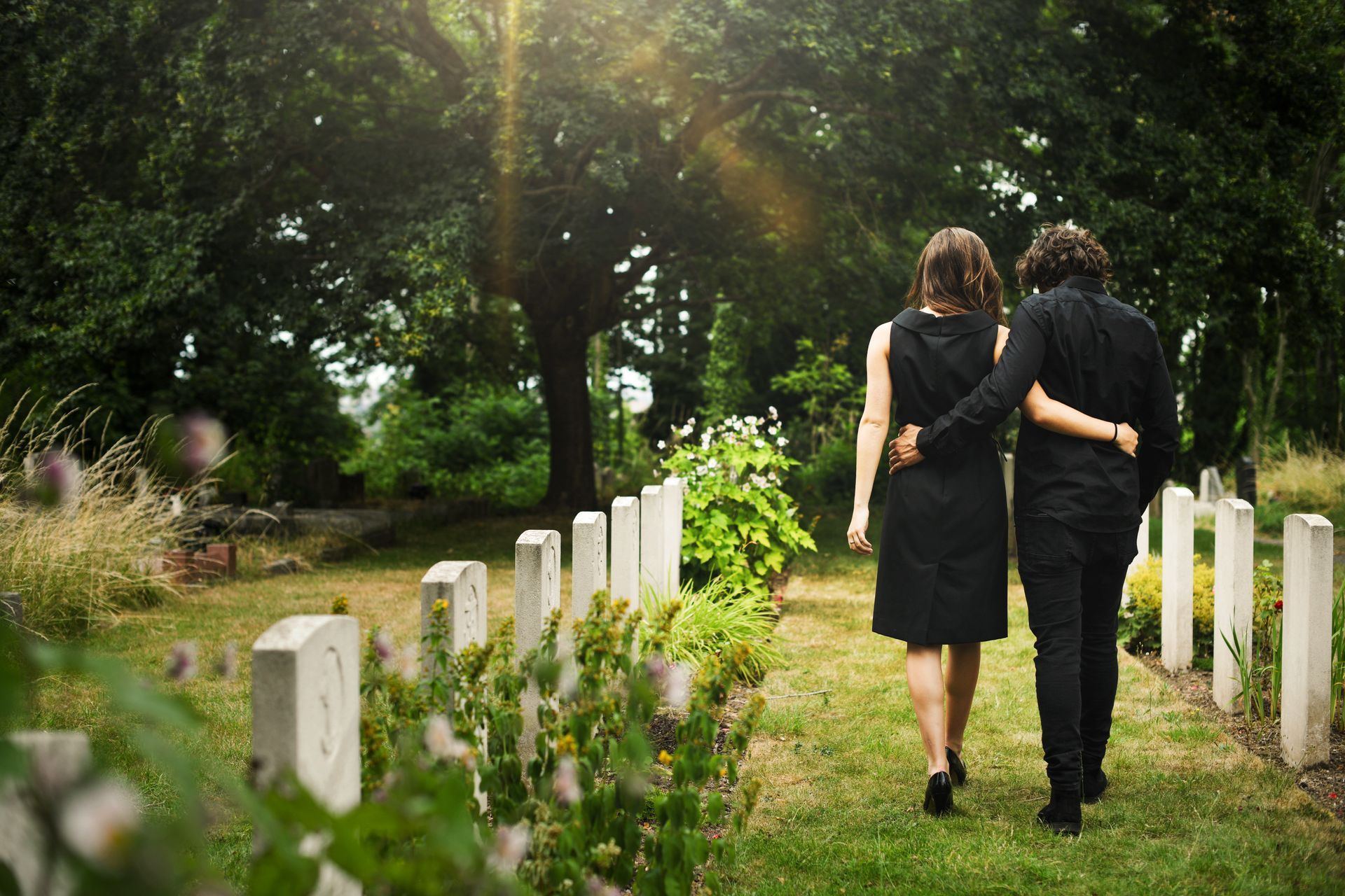 Couple in mourning walking past rows of upright monuments in a tree-lined cemetery