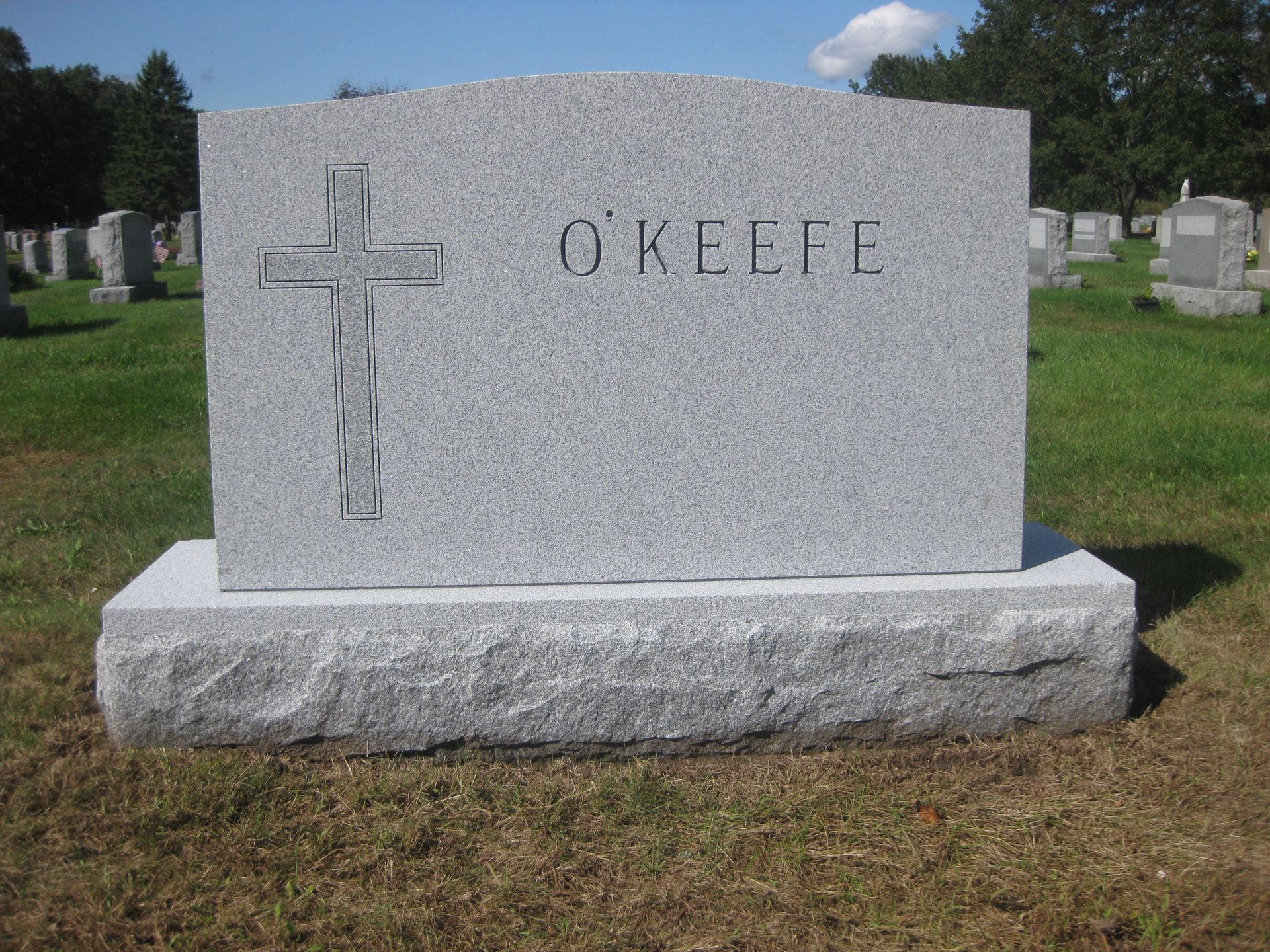 A gray granite O'KEEFE tombstone with a cross engraving, centered in a grassy cemetery.