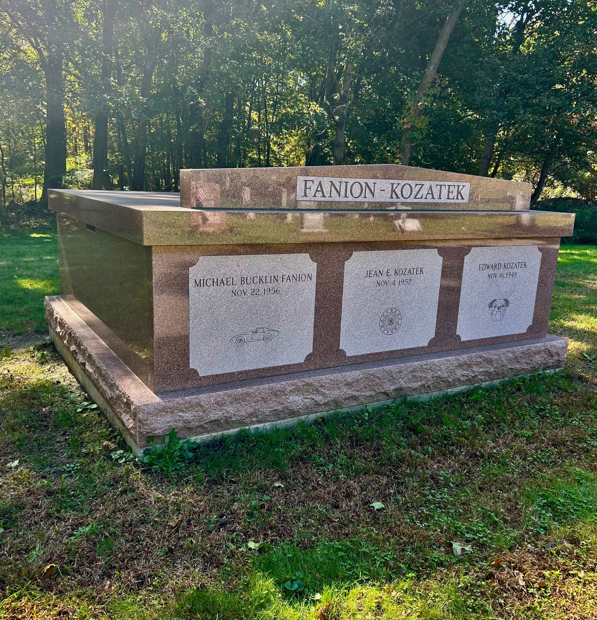 A large, rectangular granite monument with three engraved plaques, situated on a grassy lawn surrounded by trees.