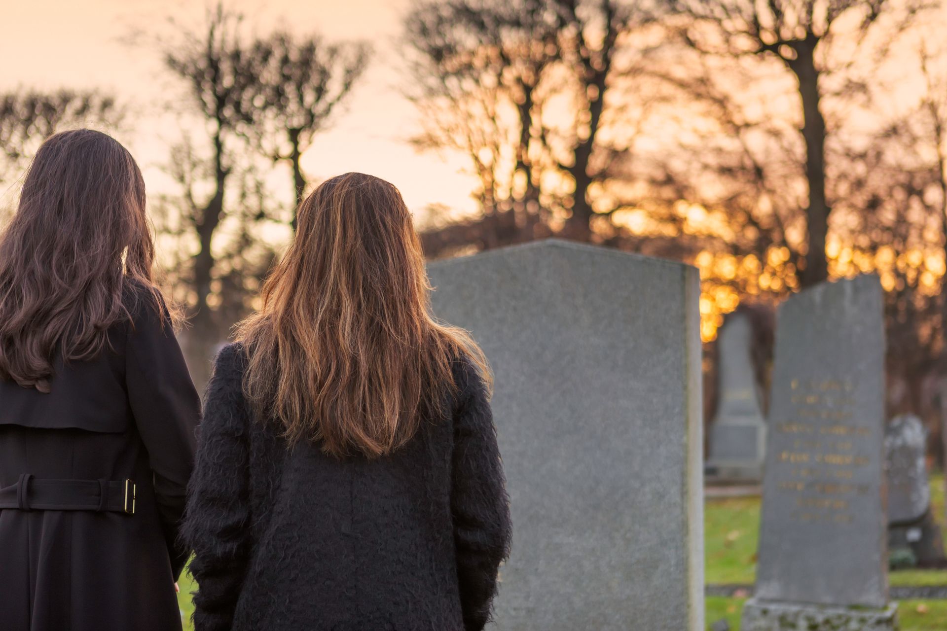 Two women in black coats at a cemetery, looking at headstones during a sunset. Marker Restoration Norton MA