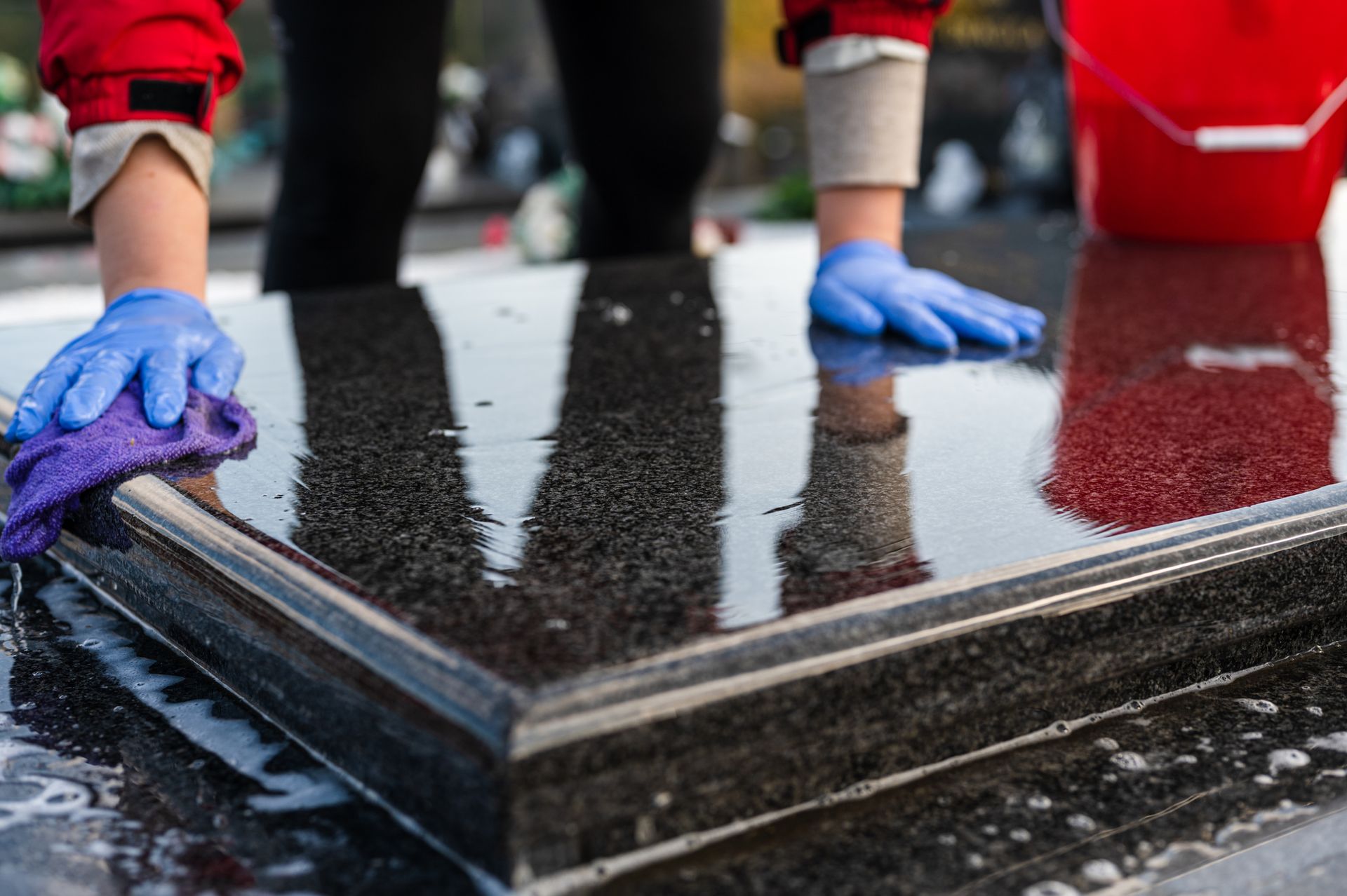 Hands in blue gloves wiping a wet, shiny black surface with a purple cloth; red bucket visible. Memorial Monuments Norton MA
