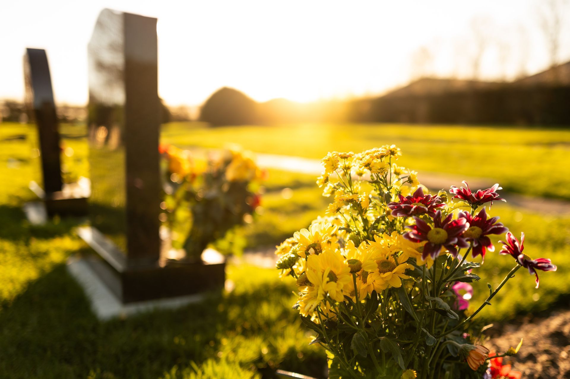 Graveyard scene, headstones with flowers in the foreground, sunlight over the green grass. Custom Monuments Restoration Norton MA