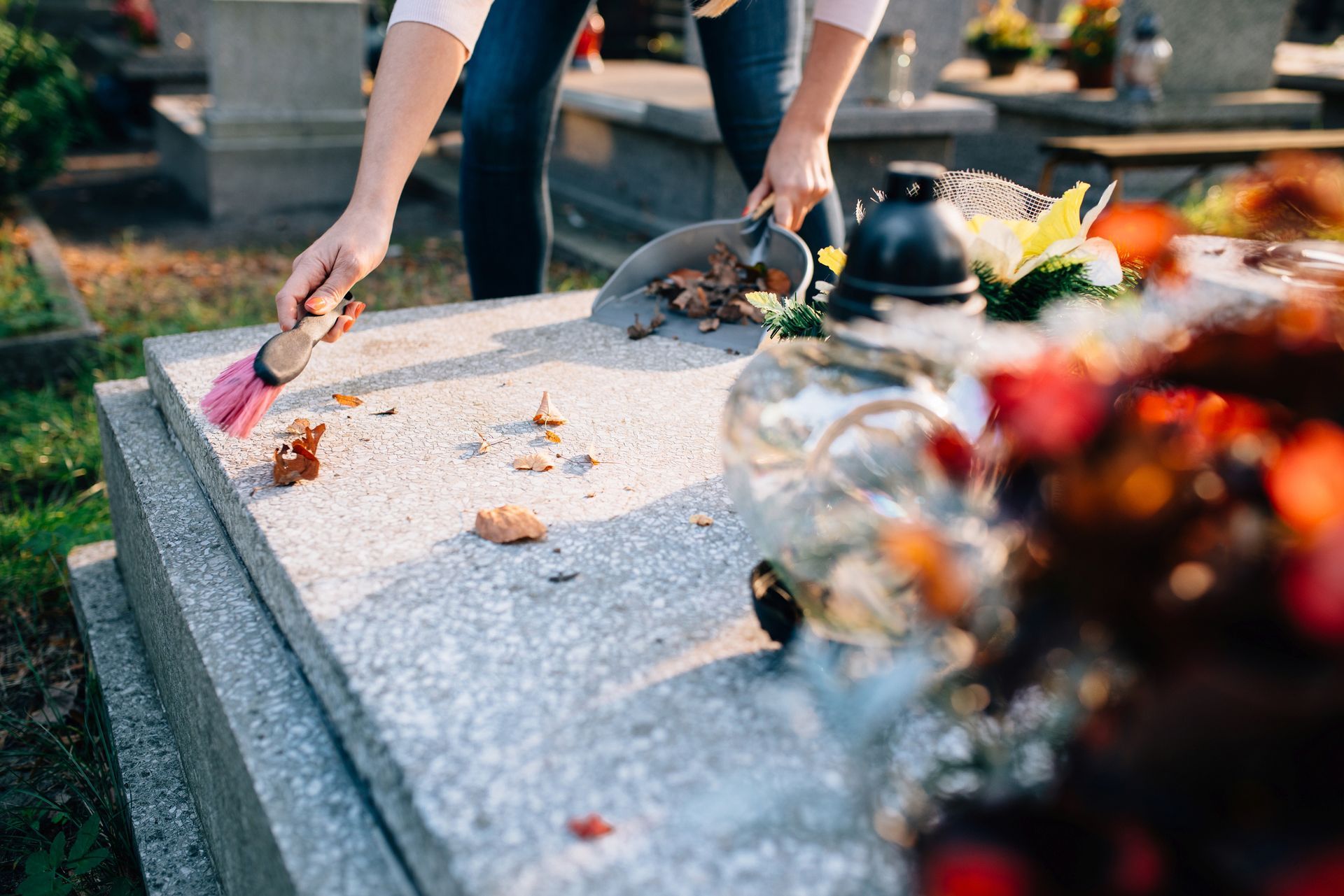 Person cleaning a tombstone with a brush and scoop in a sunlit cemetery. Monuments Restoration Norton MA