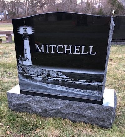 A polished black granite headstone with a laser-etched lighthouse scene and the name MITCHELL, set on a stone base.