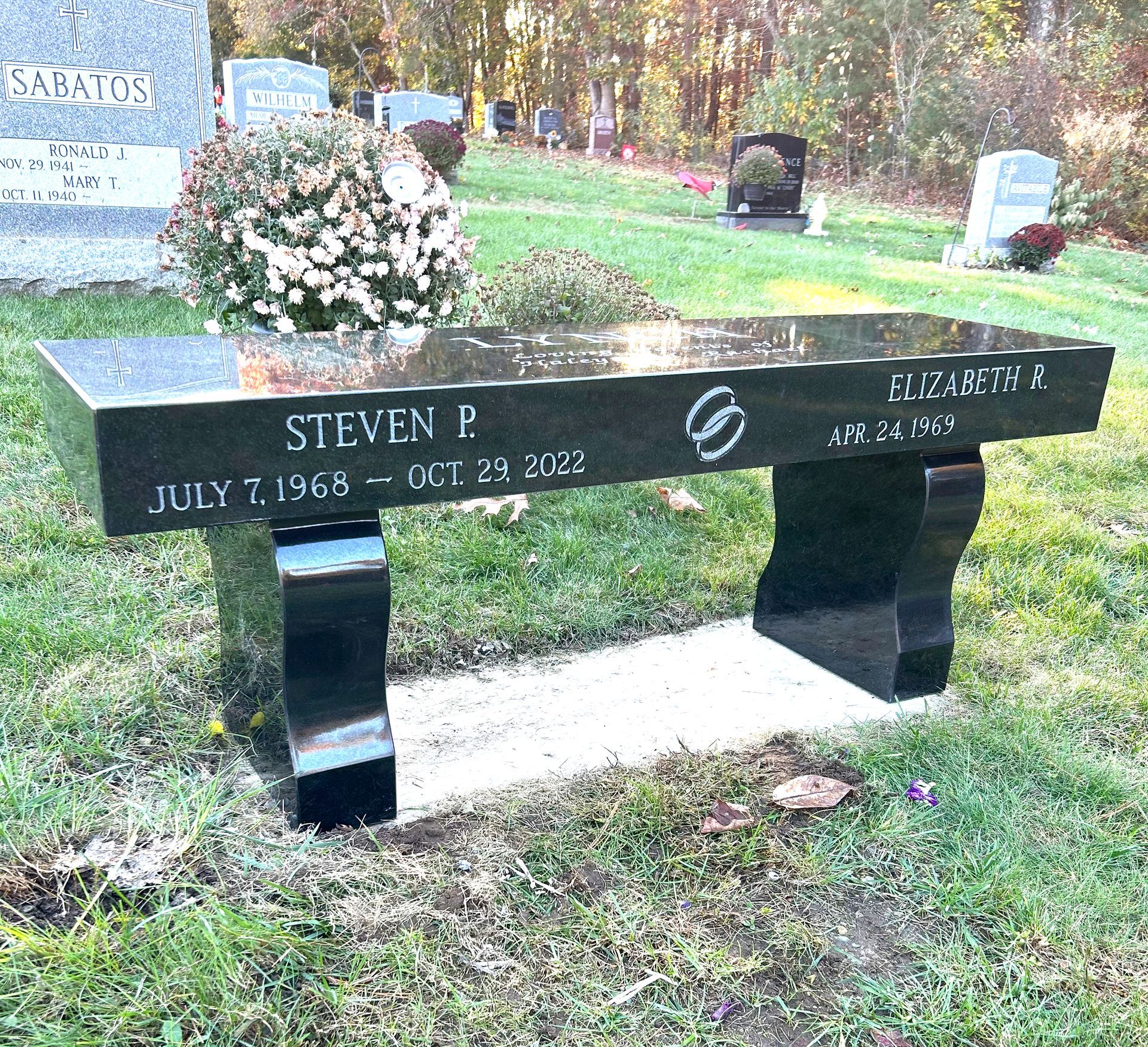 A black granite memorial bench in a cemetery inscribed with the names and dates for Steven P. and Elizabeth R.