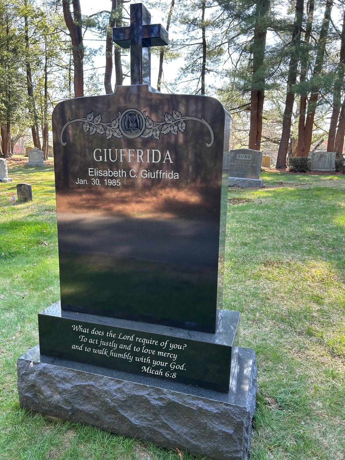 A black granite tombstone for the Giuffrida family, topped with a cross, set in a cemetery among trees.