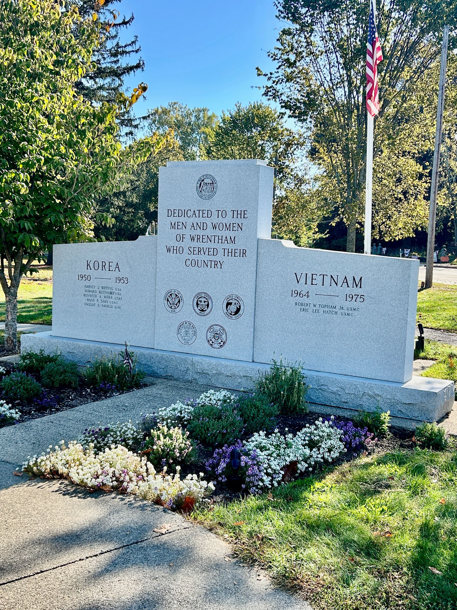 A granite veterans memorial monument in a park, featuring inscriptions for WWII, Korea, and Vietnam under an American flag.