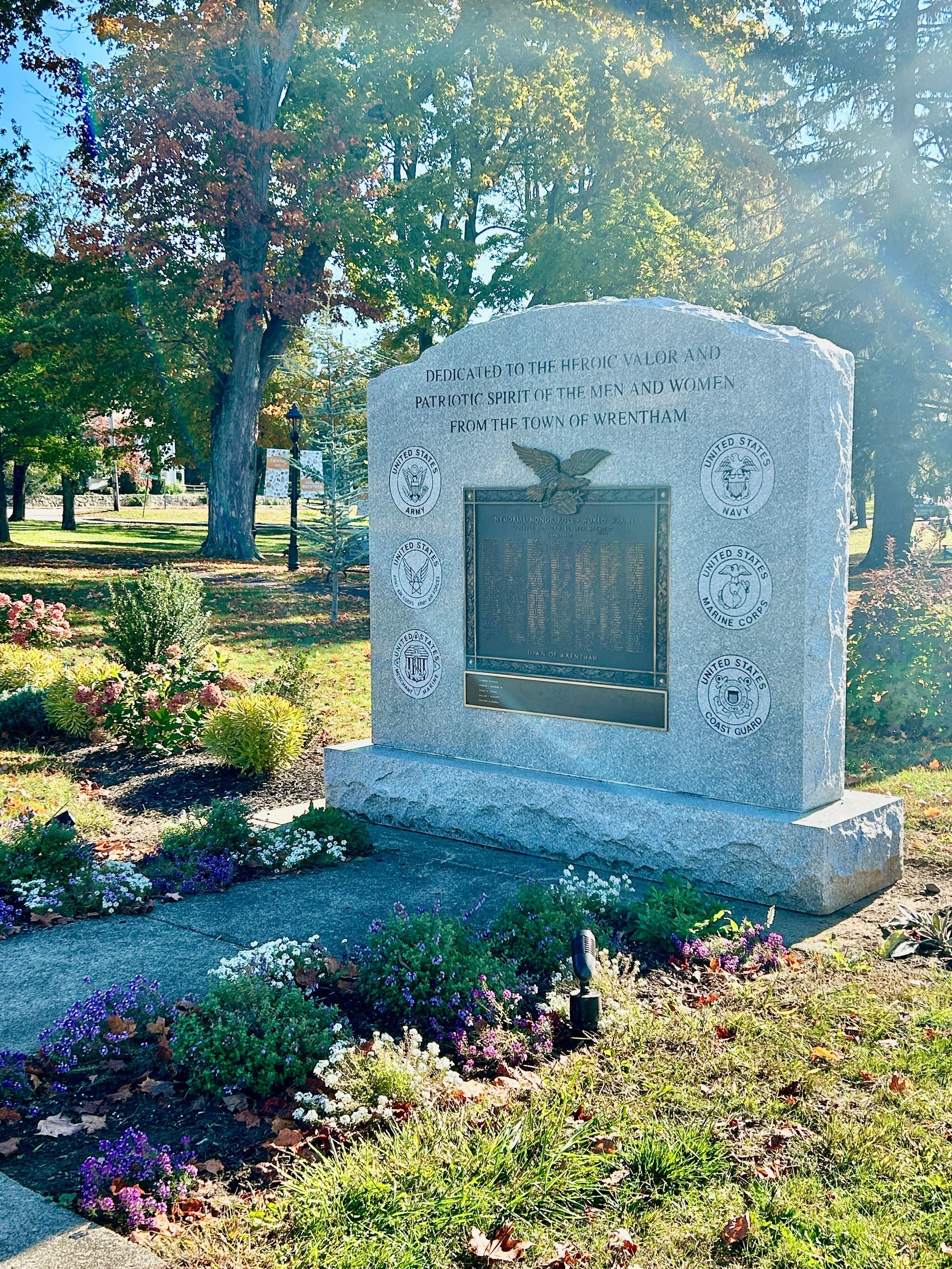 A light gray stone veterans memorial set in a park-like area with trees, flowers, and a central bronze plaque.
