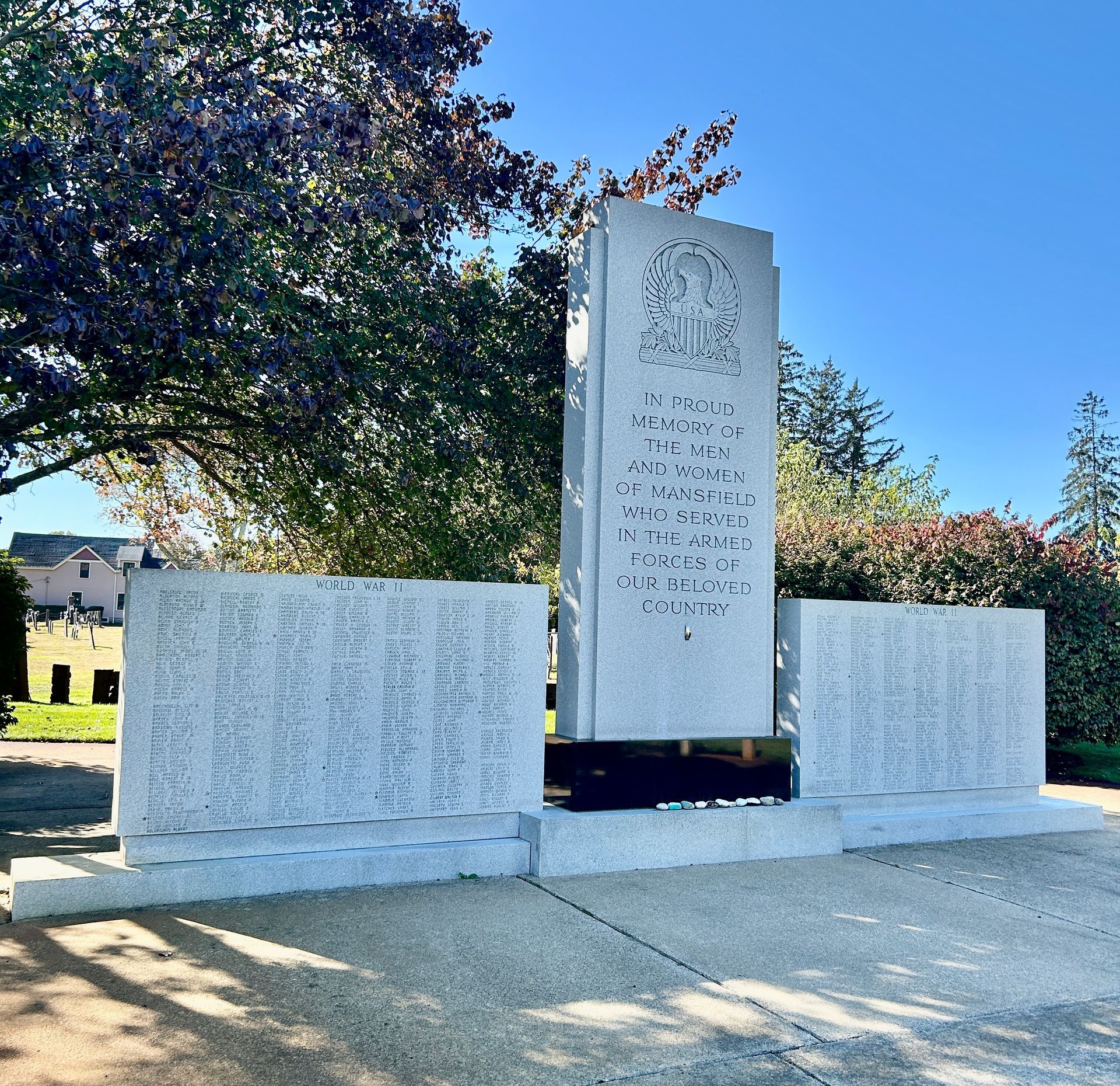 A stone war memorial with a central obelisk and two side panels engraved with names, set in a sunny, tree-lined park.