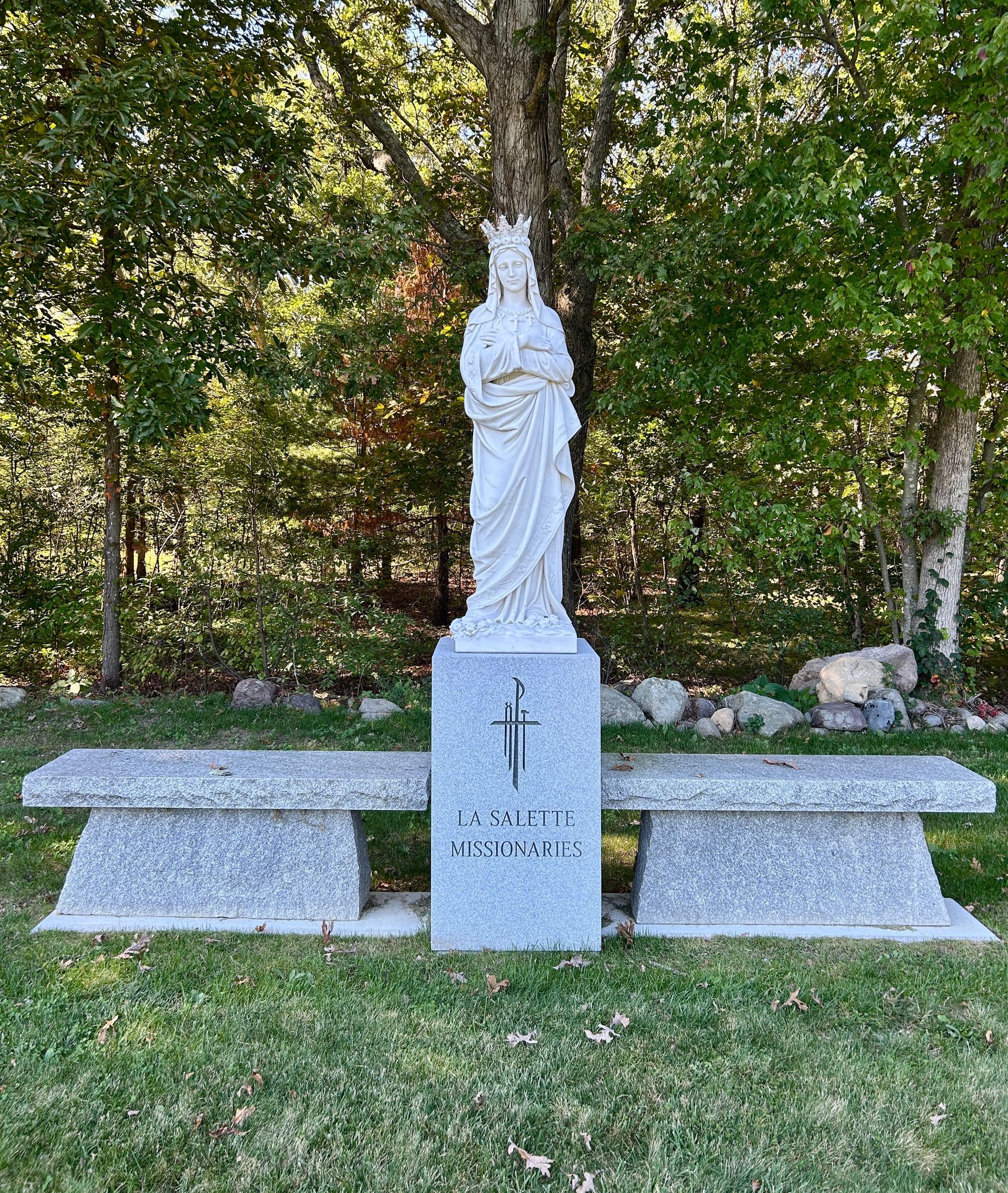 A white statue of a crowned figure stands on a gray stone plinth between two stone benches in a wooded area.