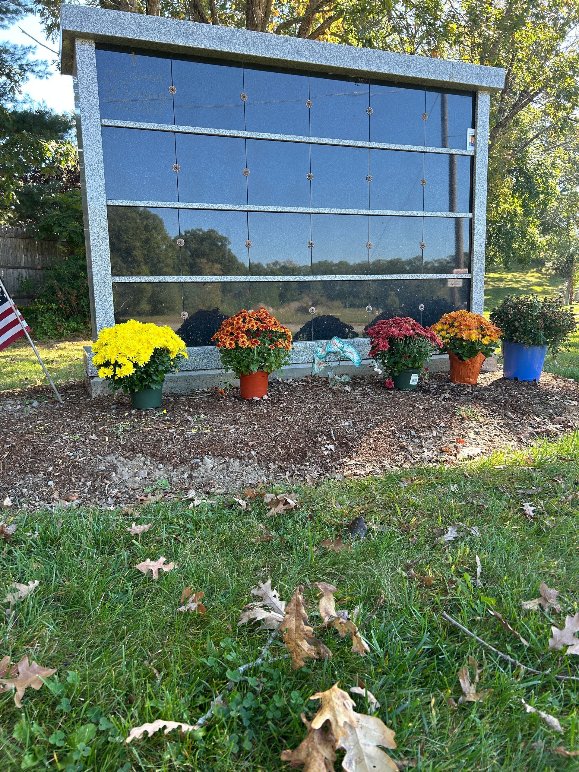 A granite columbarium with dark glass niches, decorated with several potted chrysanthemums at its base on a grassy lot.