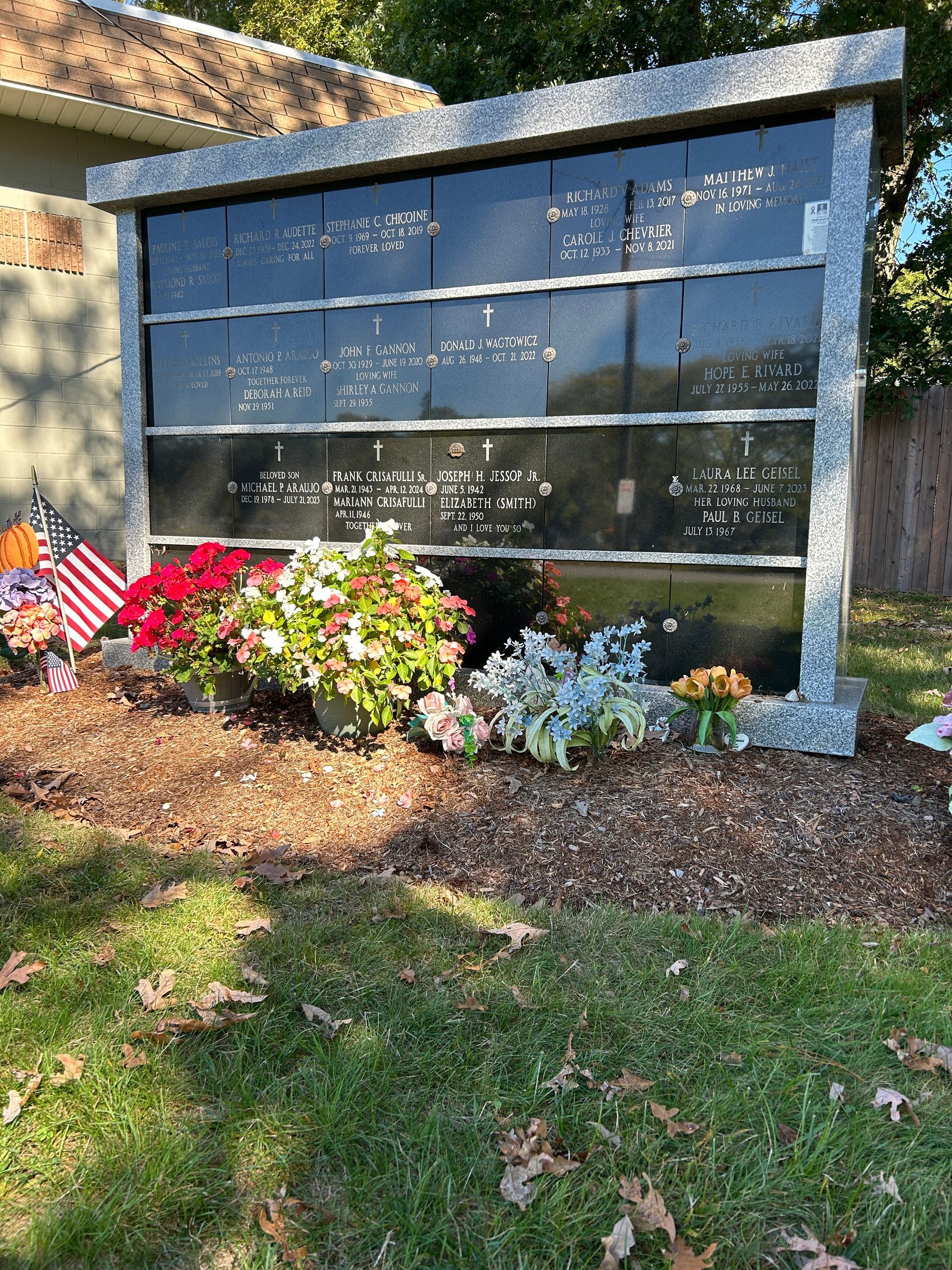 An outdoor granite columbarium with engraved memorial plaques, decorated with potted flowers and a small American flag.