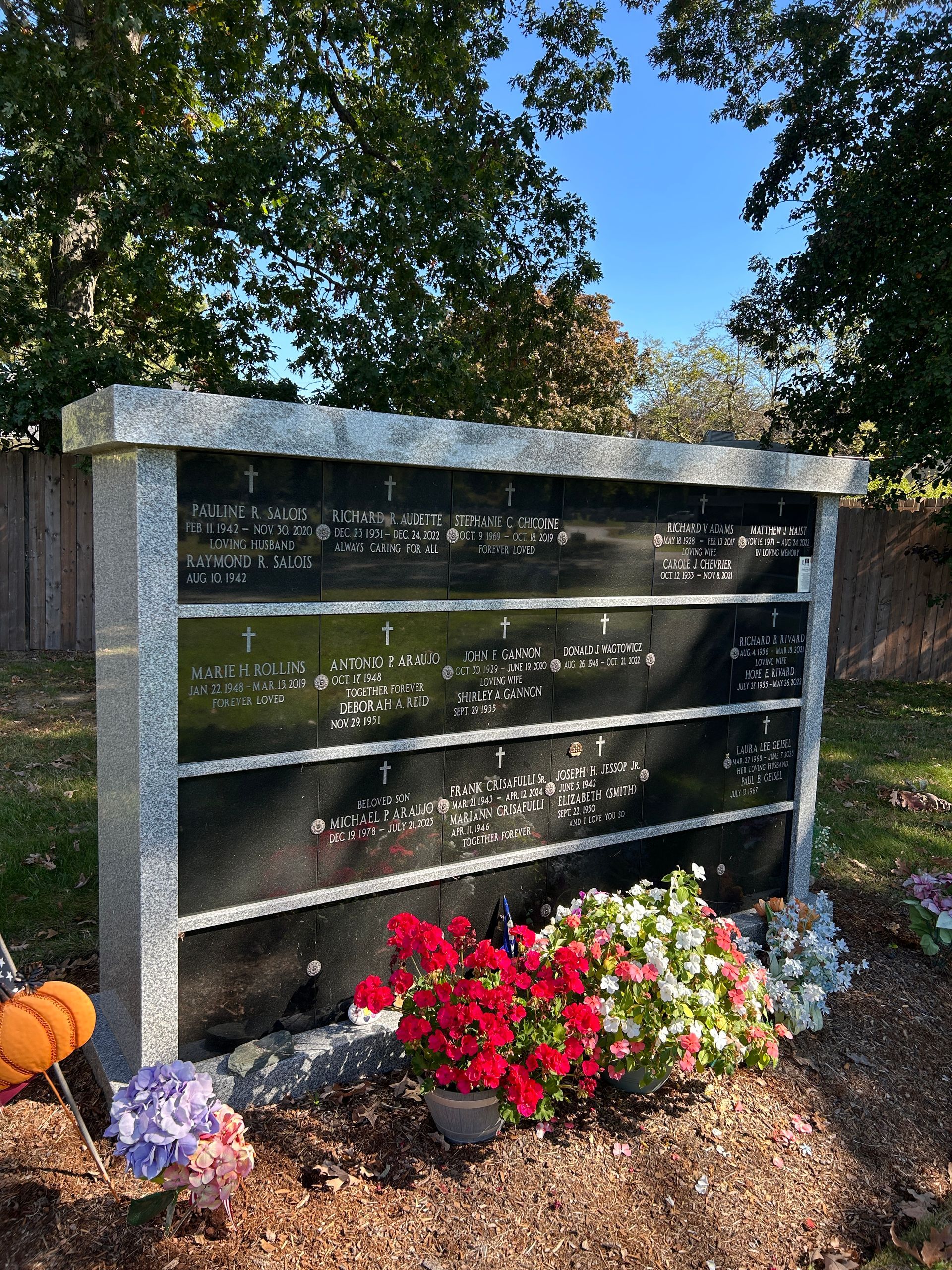 A granite columbarium with rows of memorial plaques, adorned with flowers and a small pumpkin in a cemetery.