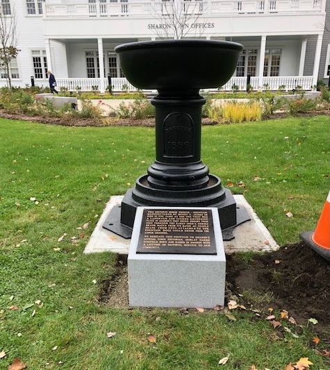 A black pedestal birdbath sits on a concrete base with a memorial plaque in front of a white building on a grassy lawn.