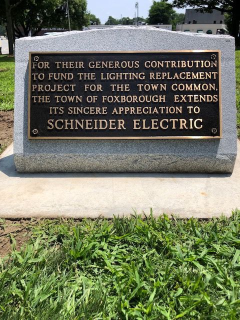 A gray stone marker with a bronze plaque thanking Schneider Electric for funding the Foxborough Town Common lighting.