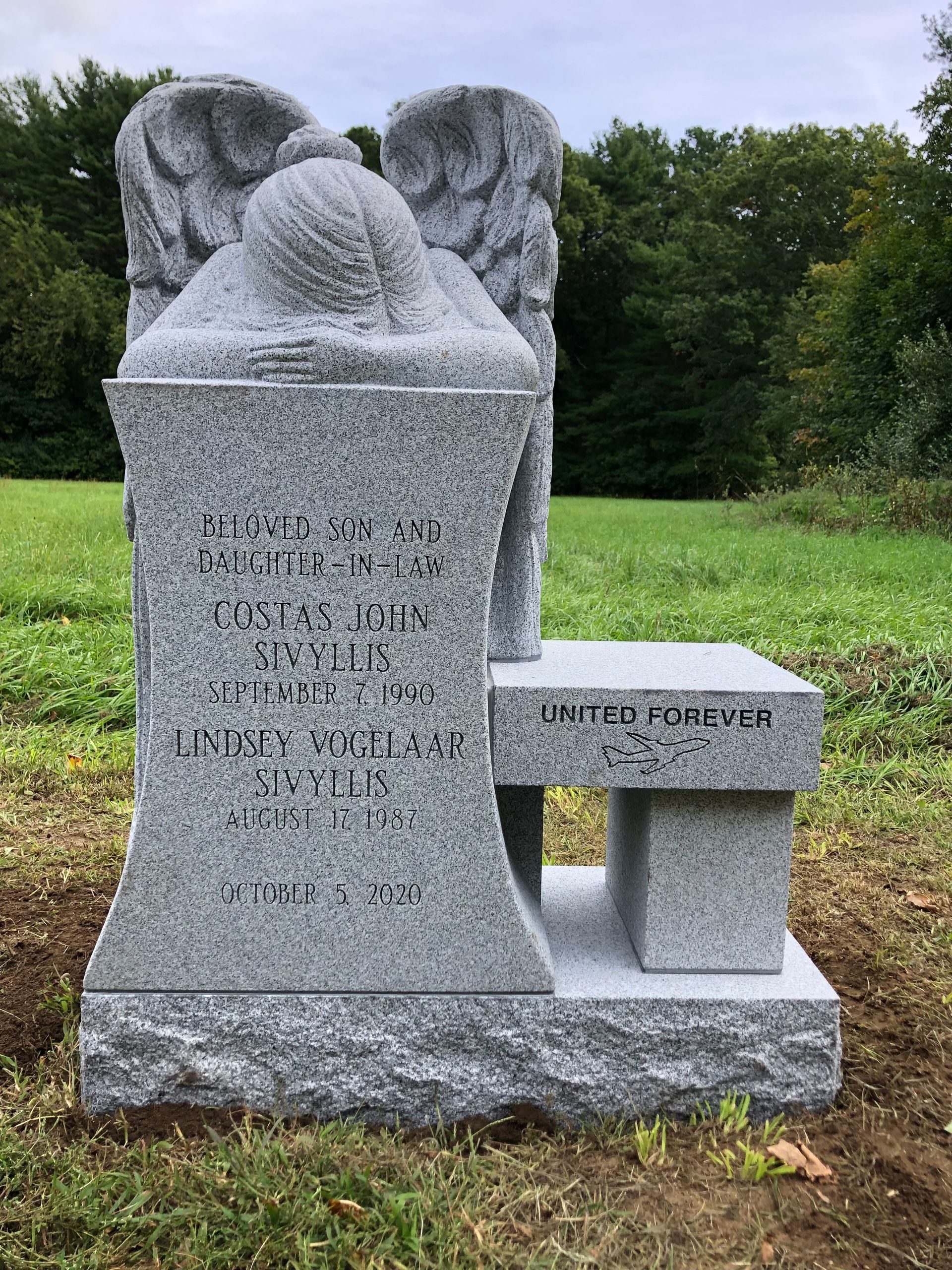 A granite headstone featuring a sculpted weeping angel, set in a grassy field with 