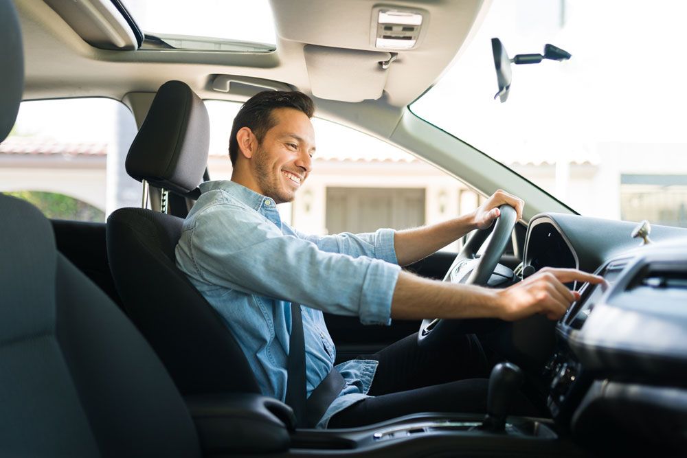 Man in a car, smiling, reaching for the dashboard controls. Sunny interior.