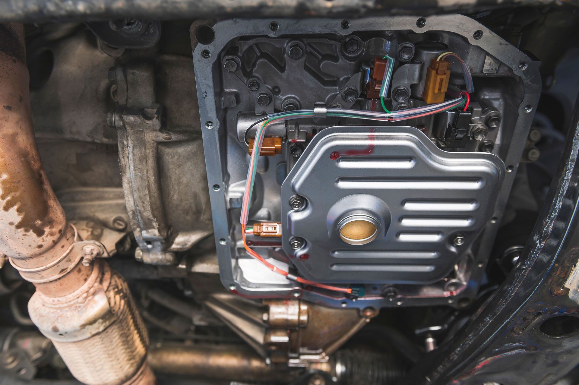 Underside of a car showing an open transmission pan with internal components and a new filter.