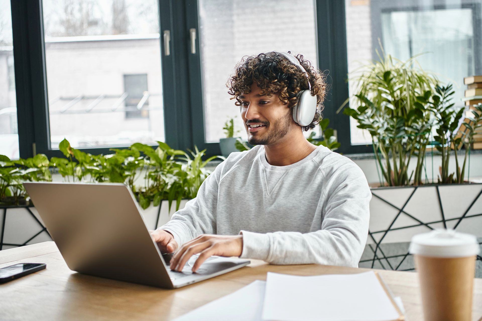 Una persona lavora al computer portatile seduta a una scrivania in un ufficio luminoso con piante e una tazza di caffè.