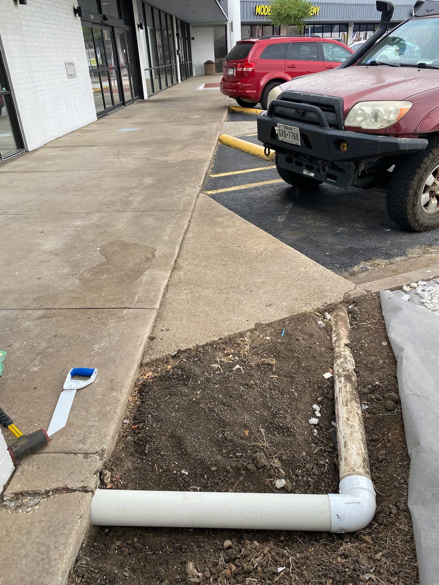 Parking lot sidewalk with a damaged white PVC pipe around a dirt patch, next to parked cars and a building.