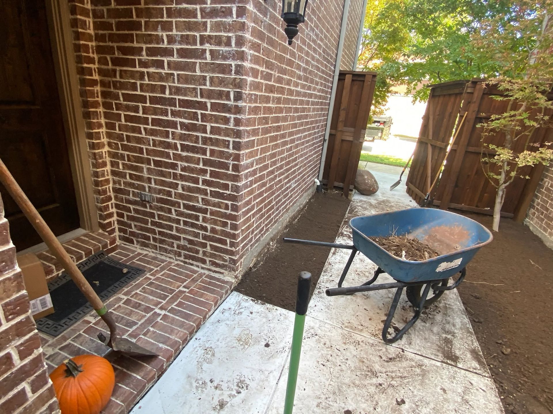 Wheelbarrow filled with leaves beside a brick house and fence, with a pumpkin on the porch.