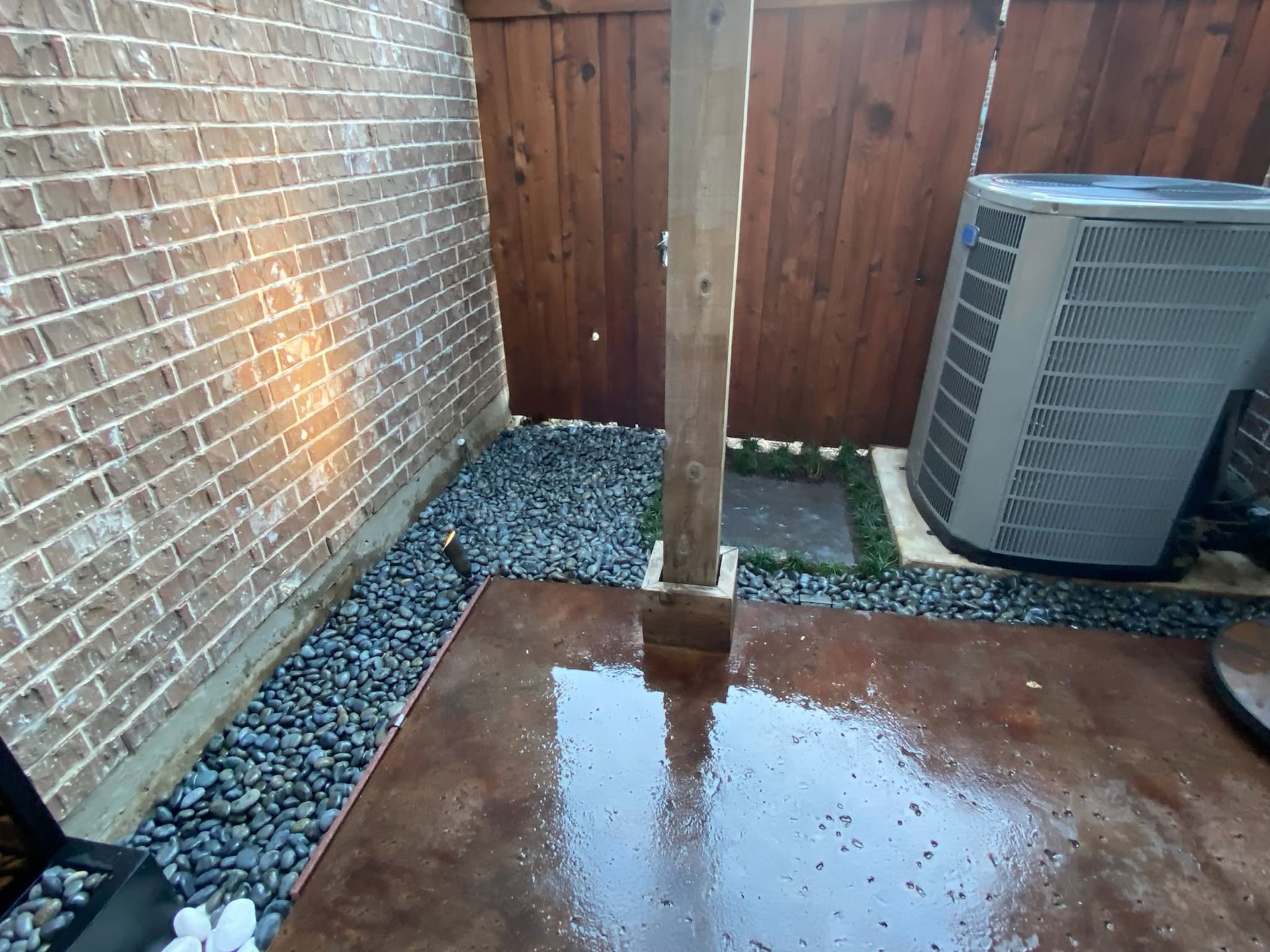 Small backyard patio with wet concrete, gravel border, brick wall, wooden fence, and an HVAC unit.