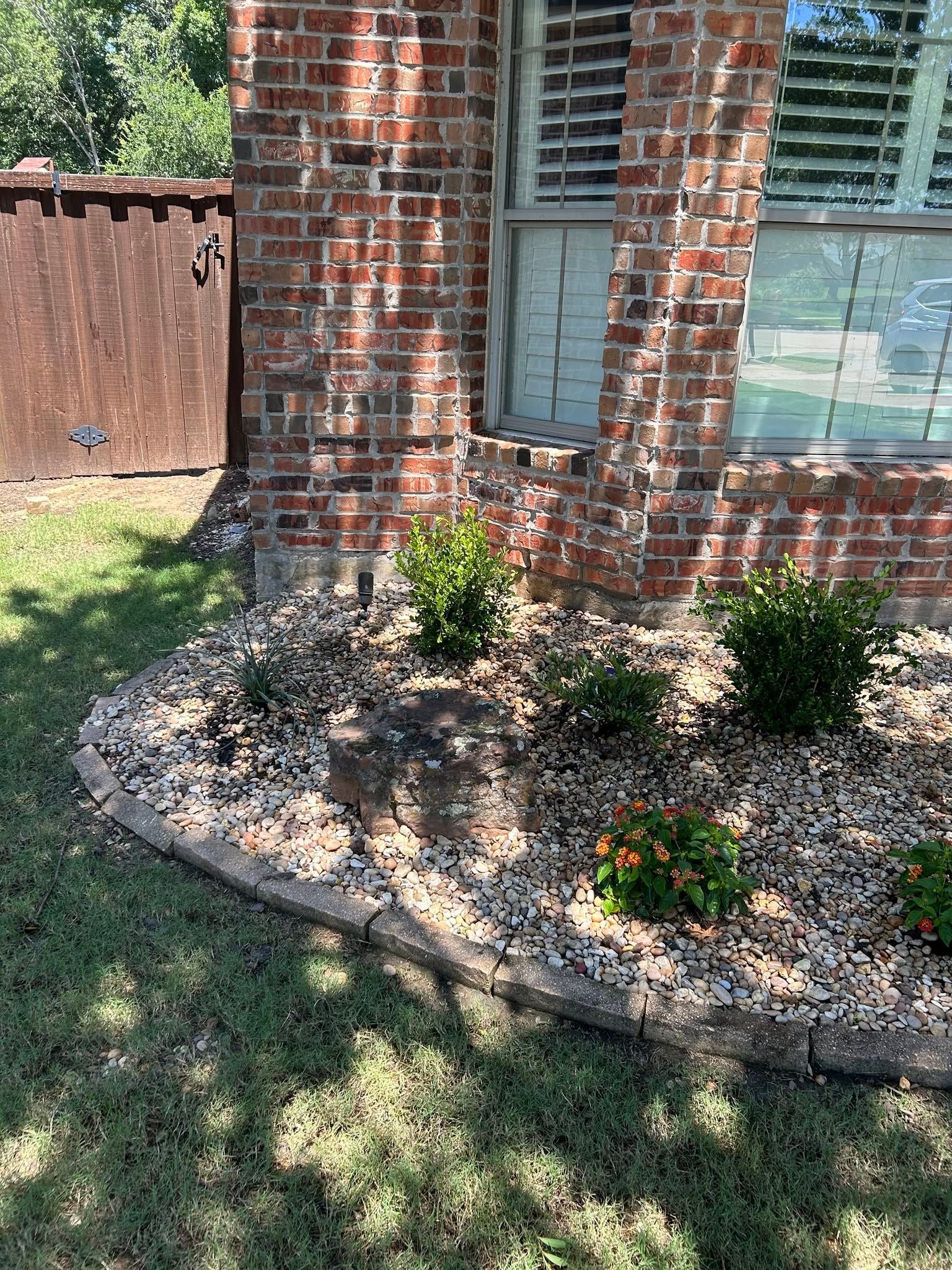 Brick house corner with a curved mulch bed, small shrubs, and orange flowers beside a grassy yard