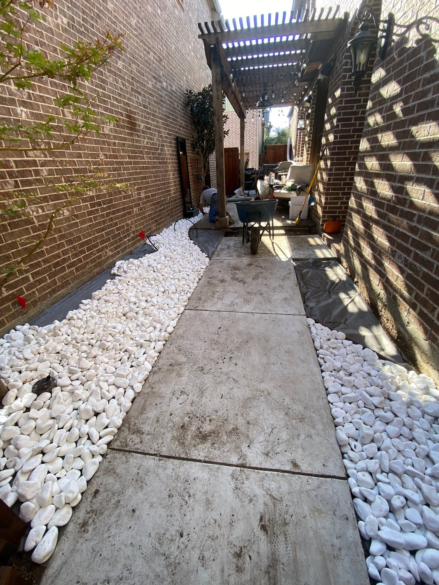 Narrow garden walkway with concrete path, white rocks, brick walls, and overhead pergola shadows