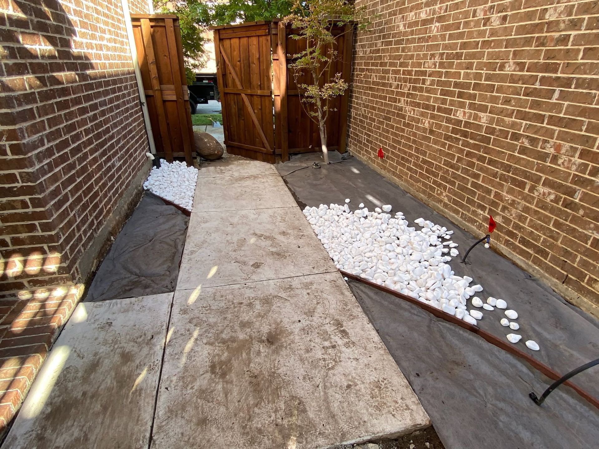 Narrow brick-lined path with a concrete walkway, wooden gate, and white gravel along both sides.