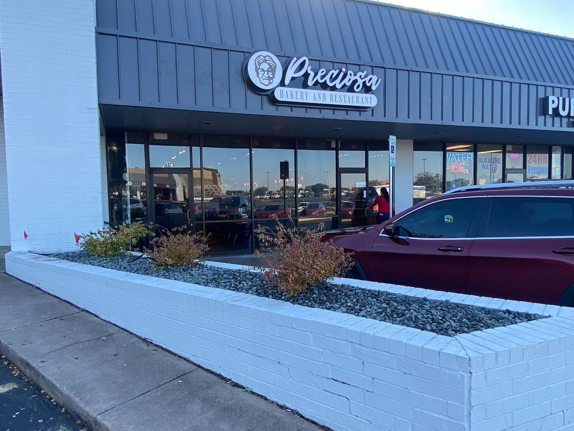 Storefront with a “Phở” sign, parked maroon car, and raised white planter in front