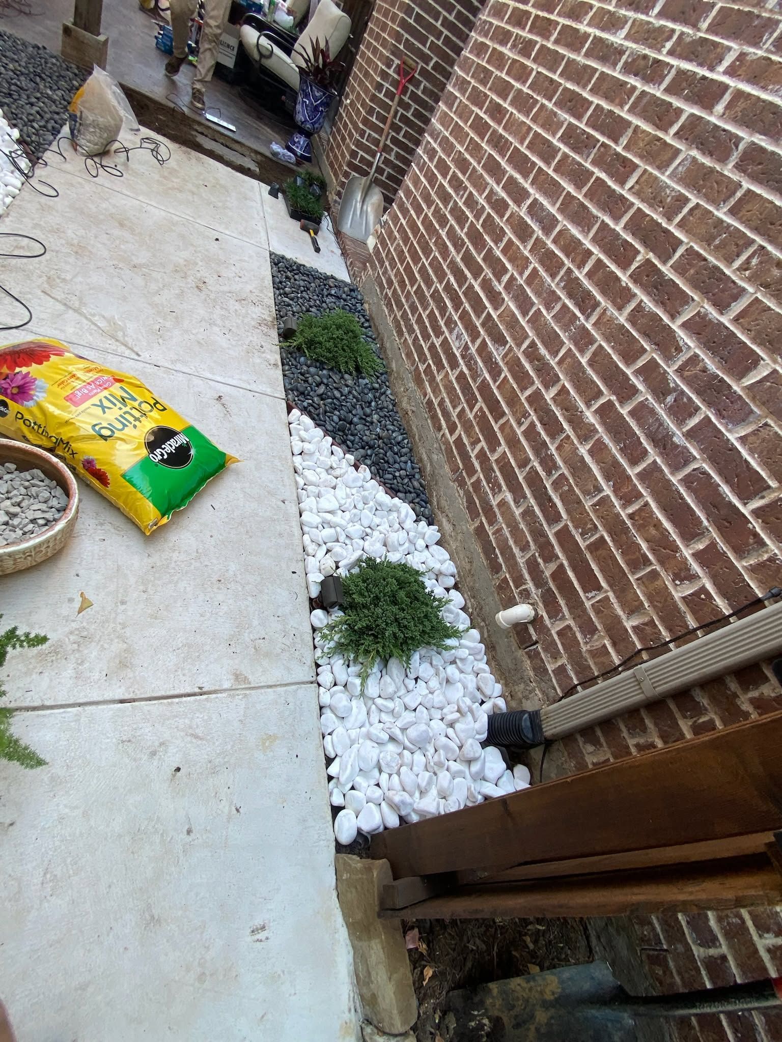 Narrow side yard with white gravel, a brick wall, concrete path, and a bag of garden mulch on the ground.