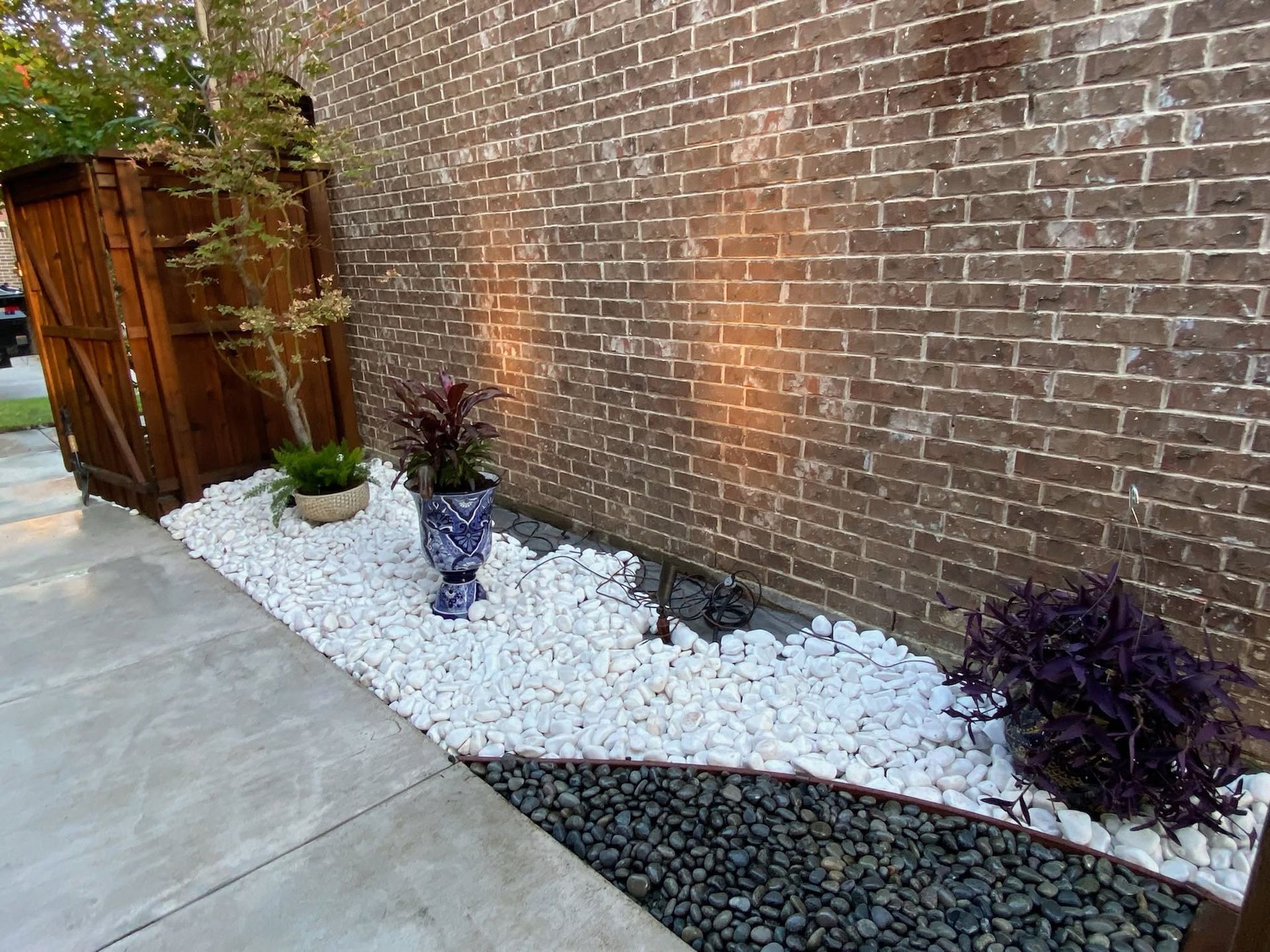 Brick wall with white and dark gravel landscaping, potted plants, and a concrete walkway along the side of a building
