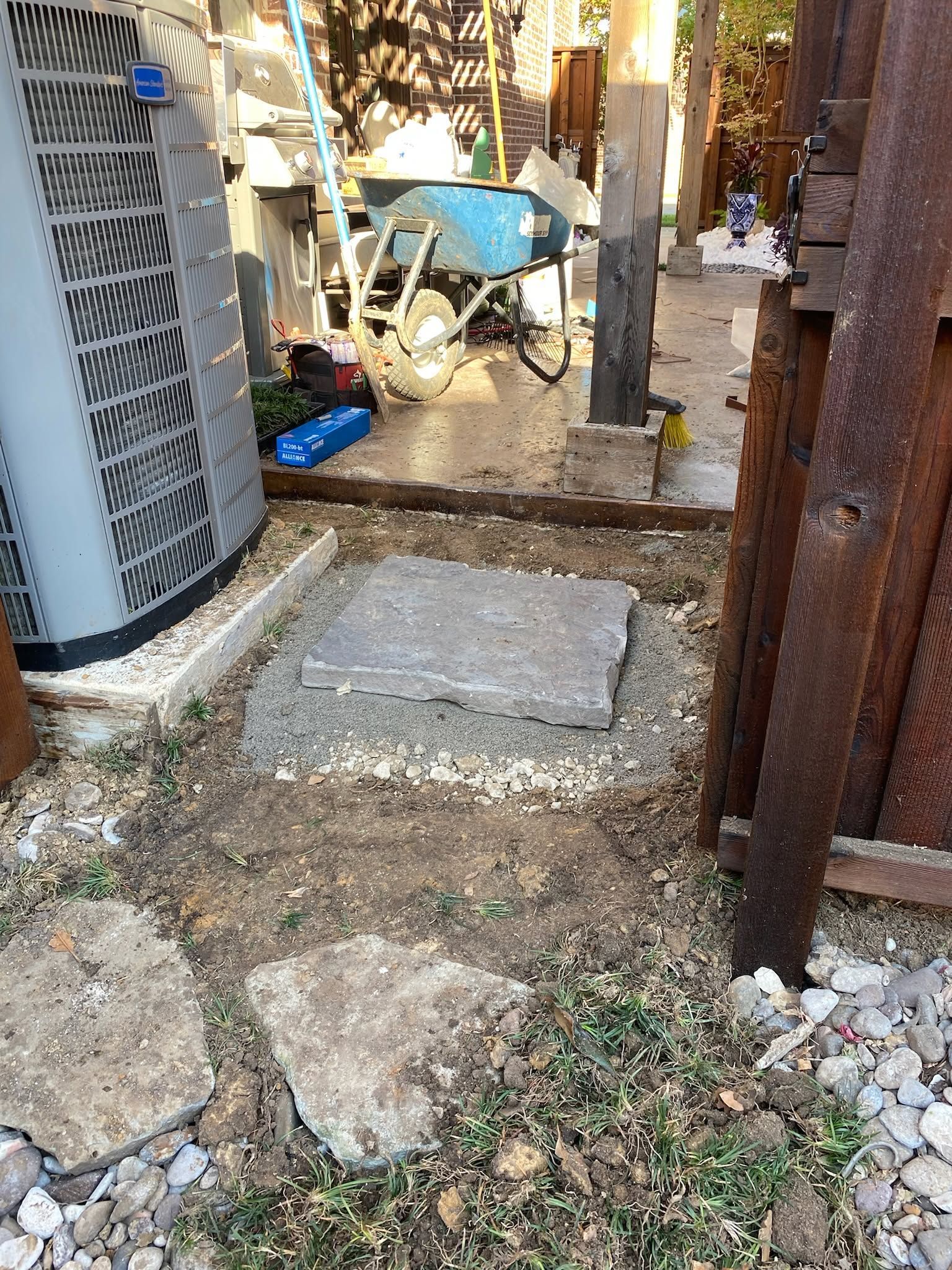 Narrow outdoor side yard with stepping stones, air conditioner unit, blue wheelbarrow, and rusty fence.