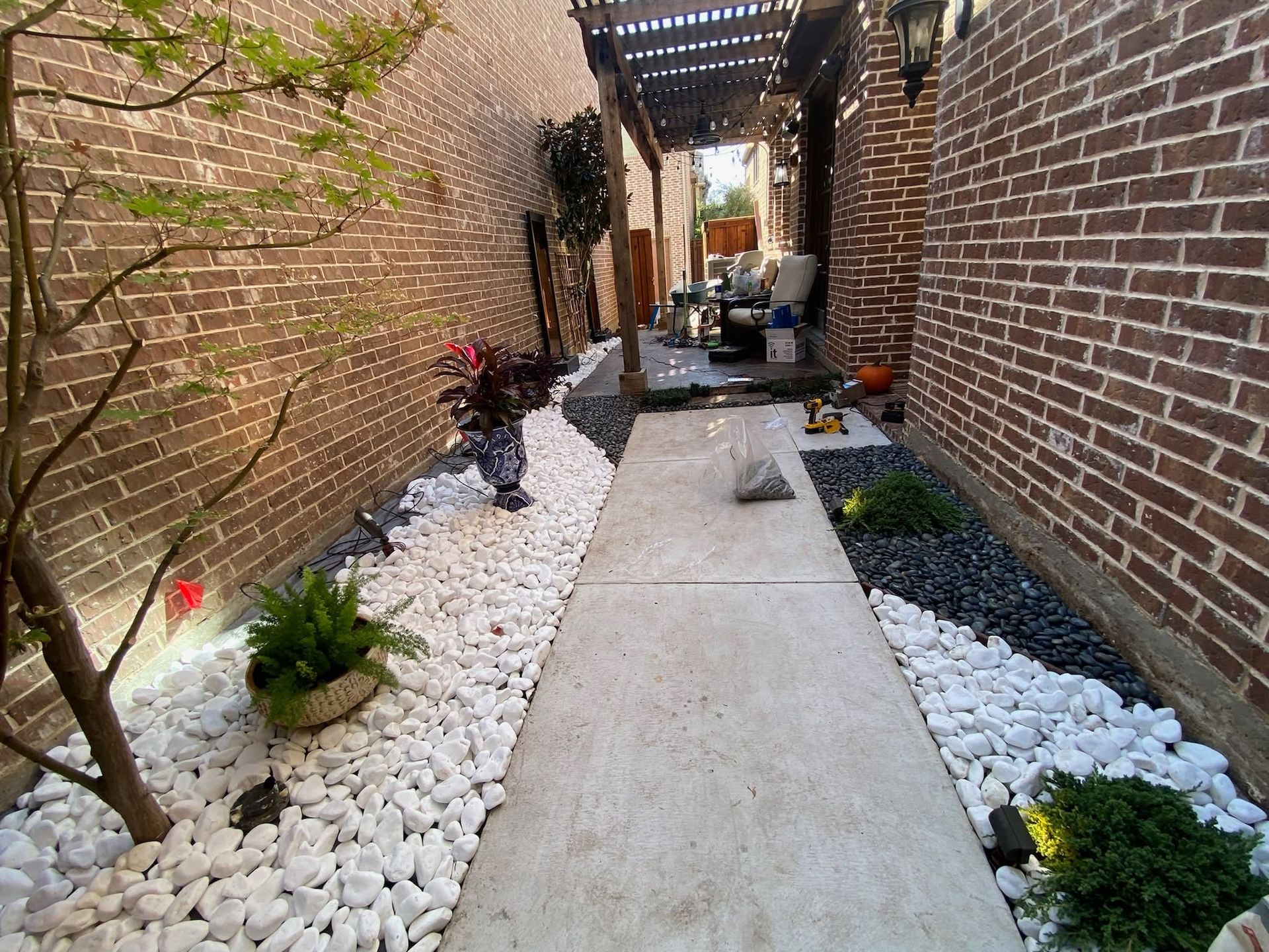 Narrow brick walkway with white gravel, potted plants, and a pergola overhead