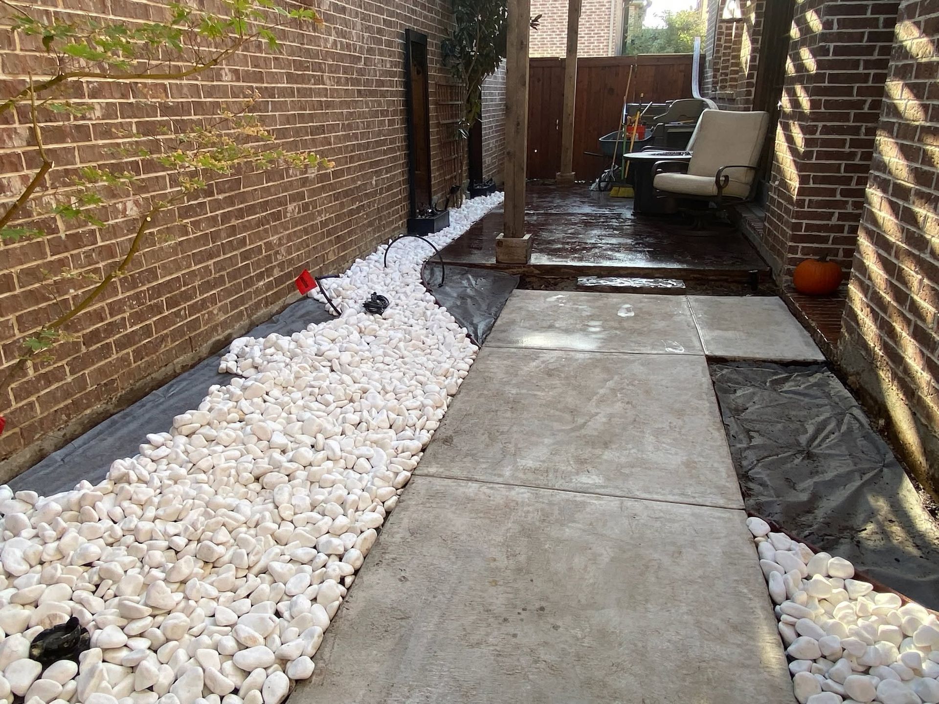 Narrow side yard with concrete path, white rocks, brick walls, and patio chairs at the far end