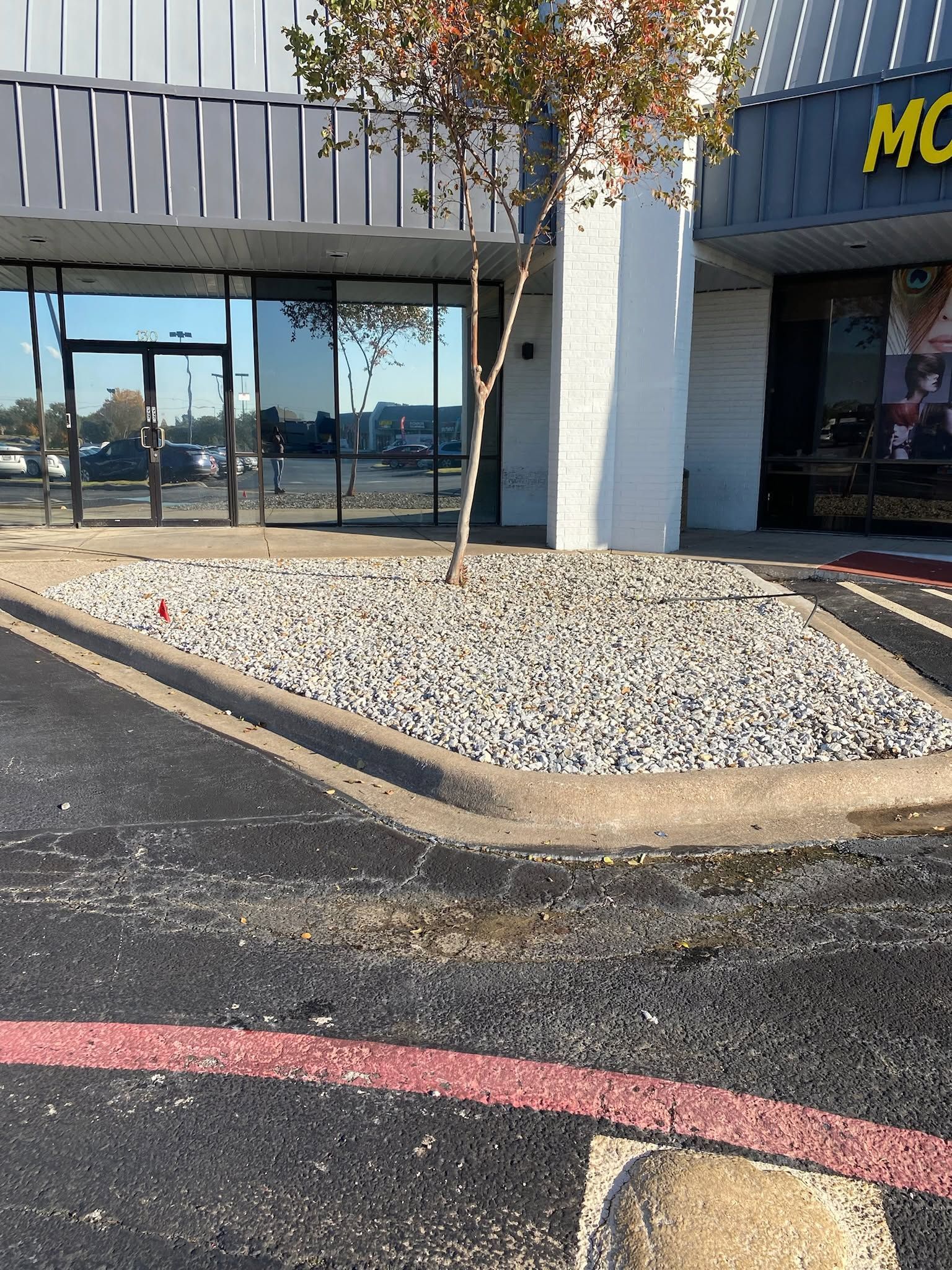 Storefront entrance with glass doors, white columns, and a rock-covered planter beside a parking lot.