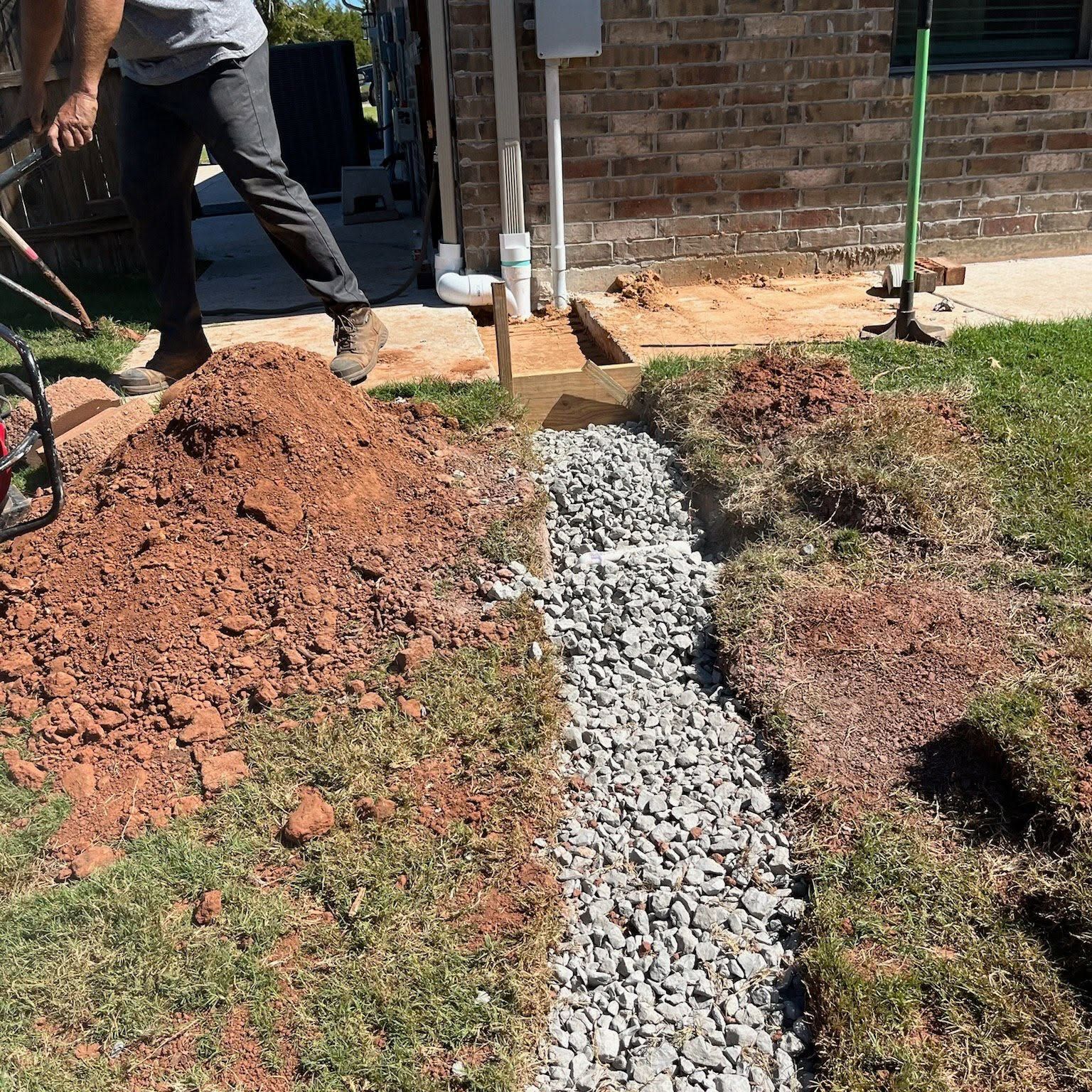 Worker spreading gravel in a narrow trench beside a house, with piles of dirt and grass nearby