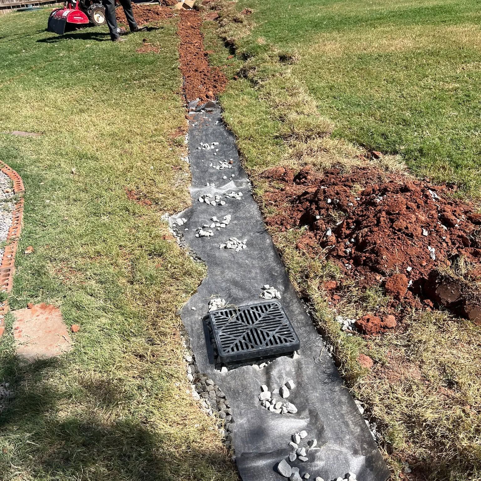 A narrow trench in a grassy yard with black pipe and a drain grate, alongside piles of excavated dirt.
