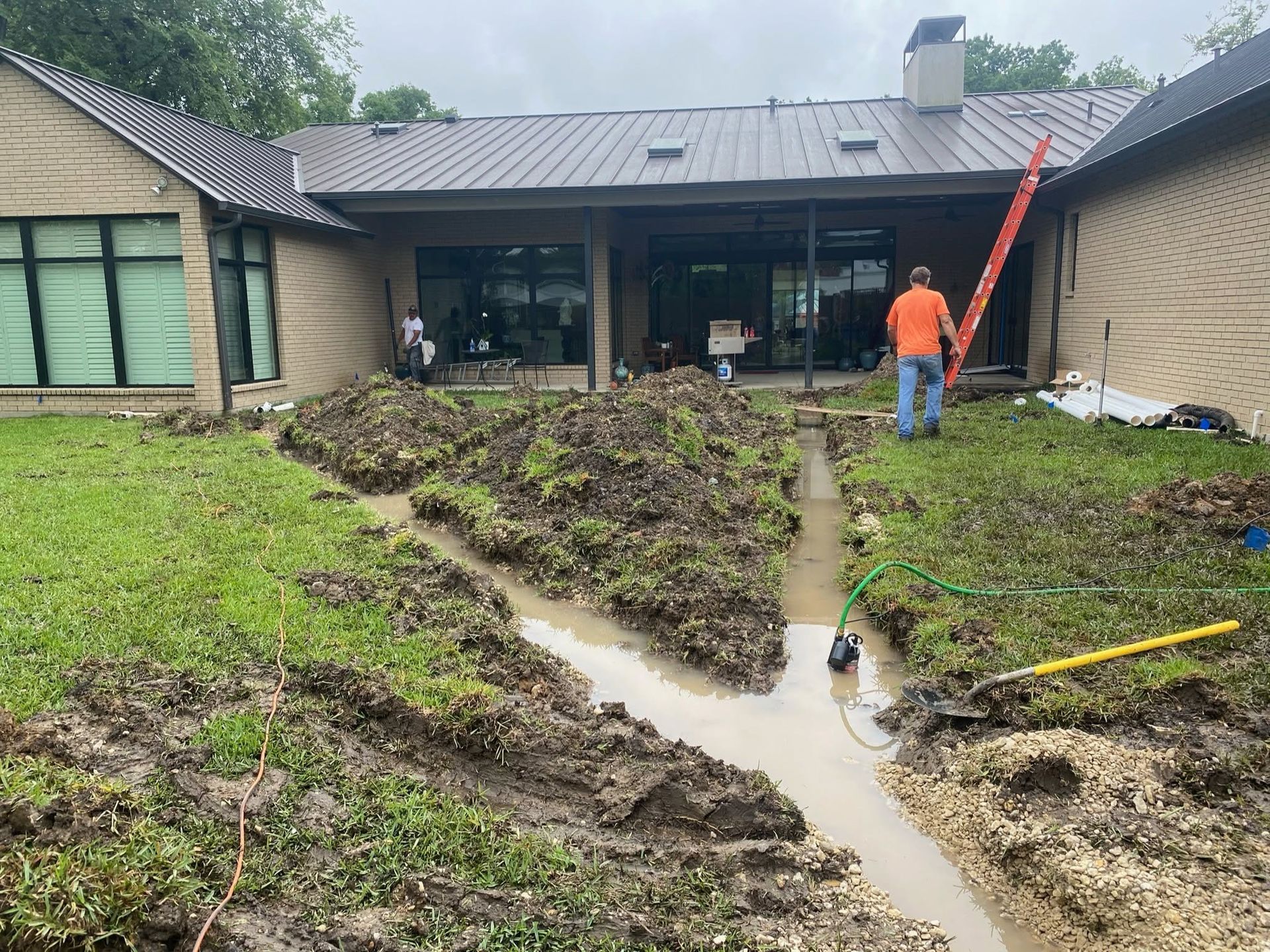 Flooded muddy yard beside a house, with a worker in orange near a ladder and damaged lawn.