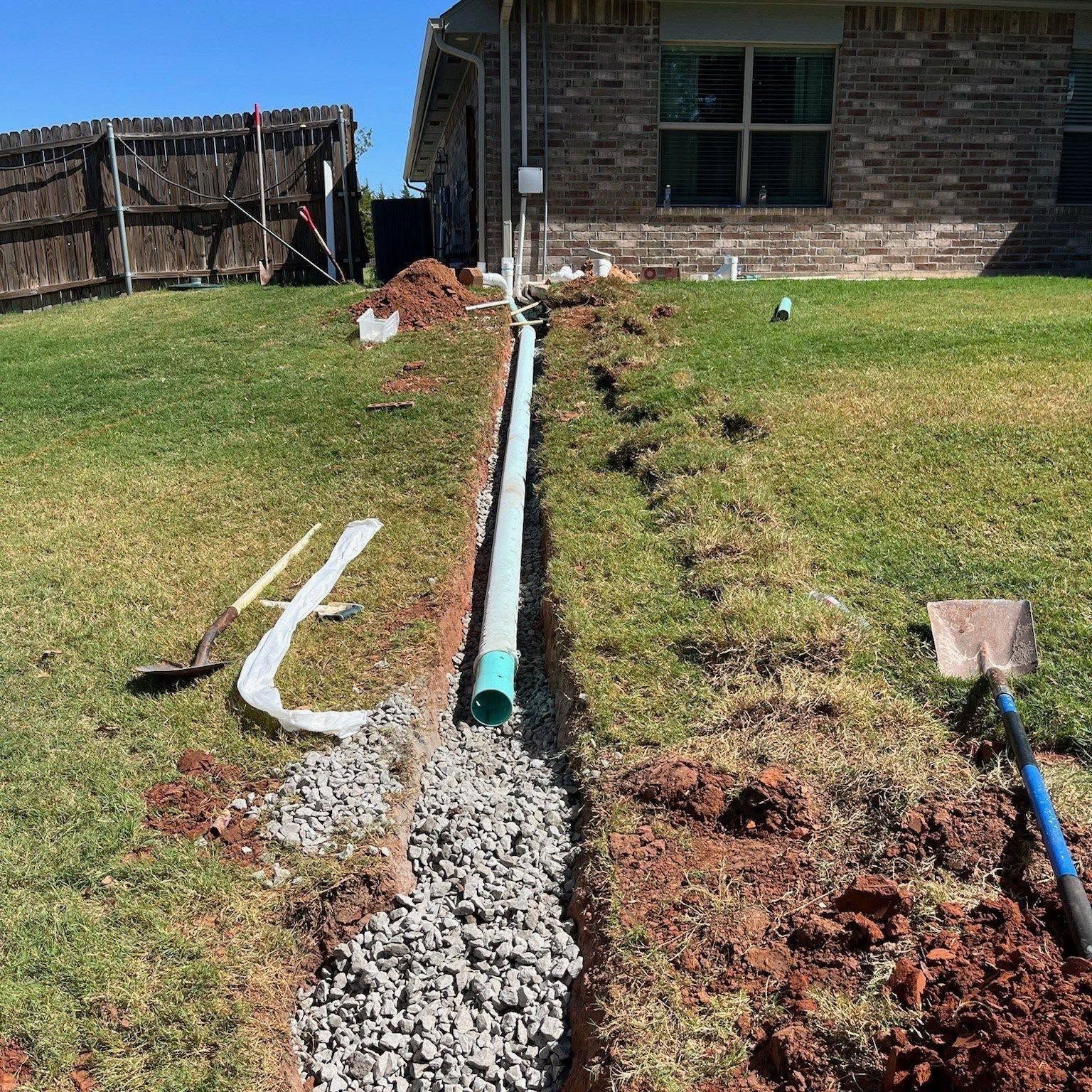 Backyard trench with gravel drain and green pipe beside a brick house, tools lying on the grass