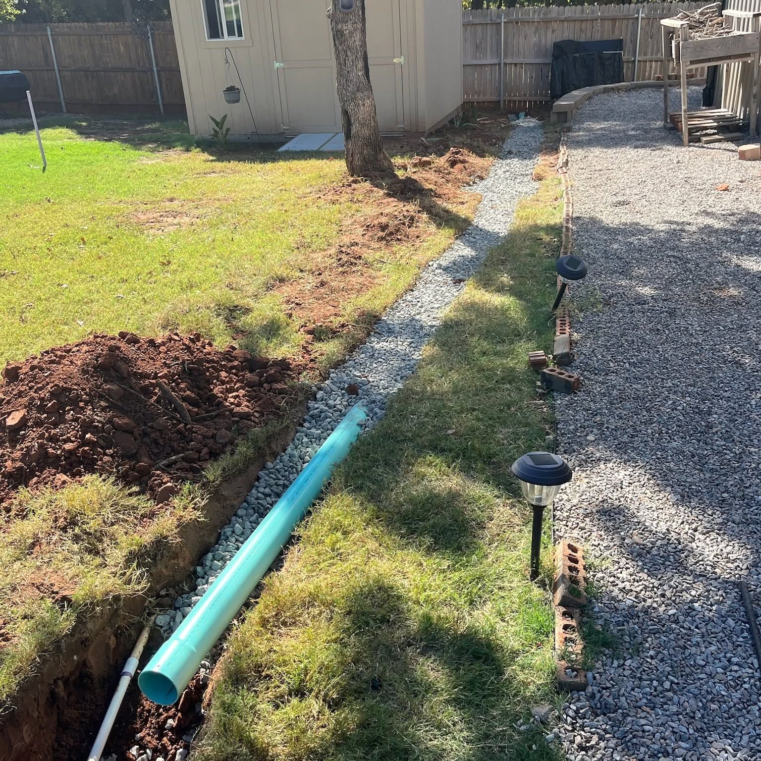 Backyard trench with a blue pipe beside grass, gravel, and small solar lights under construction.