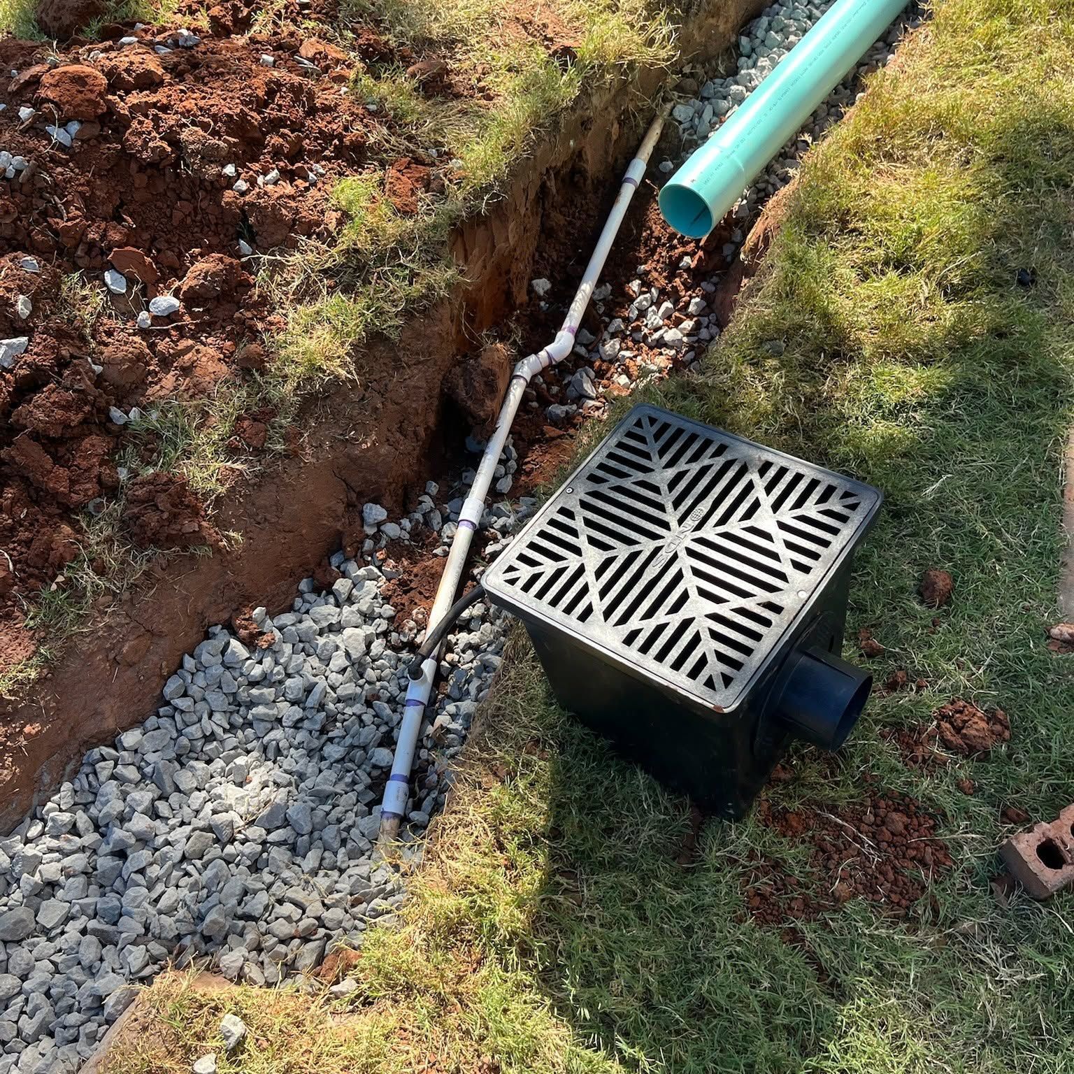 Drainage trench with gravel, black grate, and green pipe beside a grassy yard