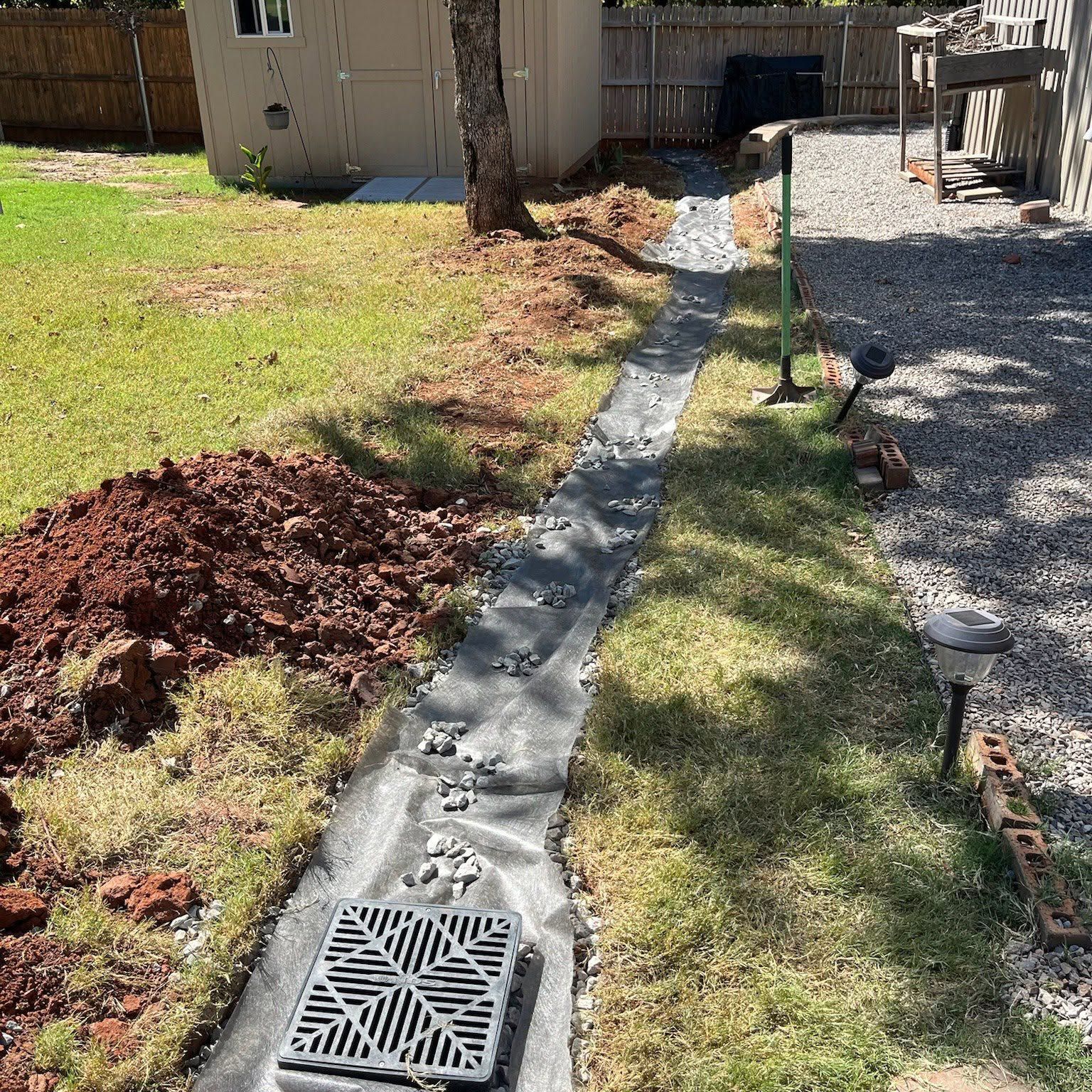 Backyard drainage trench with black liner, mulch piles, and gravel path beside a house and fence