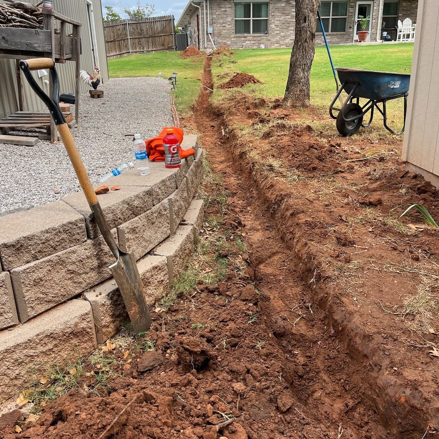 Trench dug along a backyard patio beside a house, with a shovel and wheelbarrow nearby.