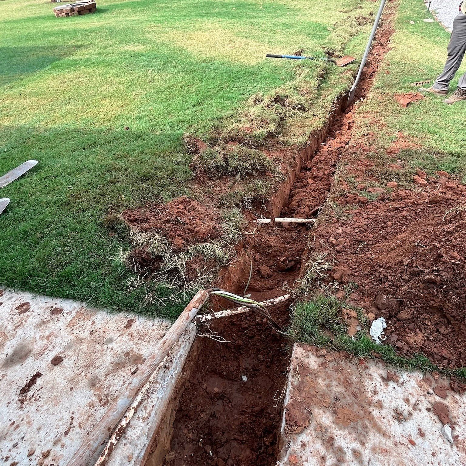 Deep trench cut through a green lawn with exposed soil and broken concrete edges