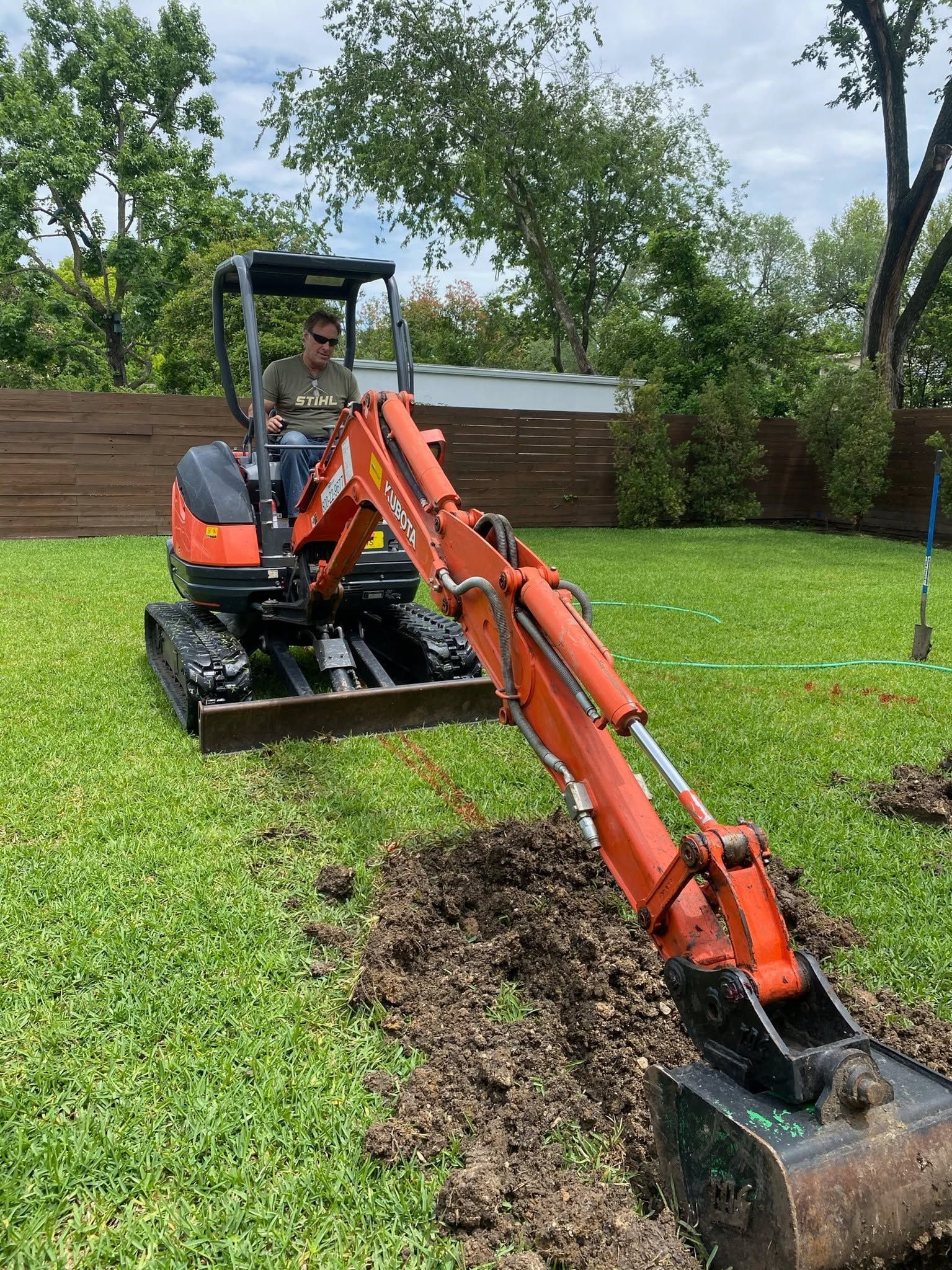 Orange mini excavator digging a trench in a grassy backyard with trees and a wooden fence