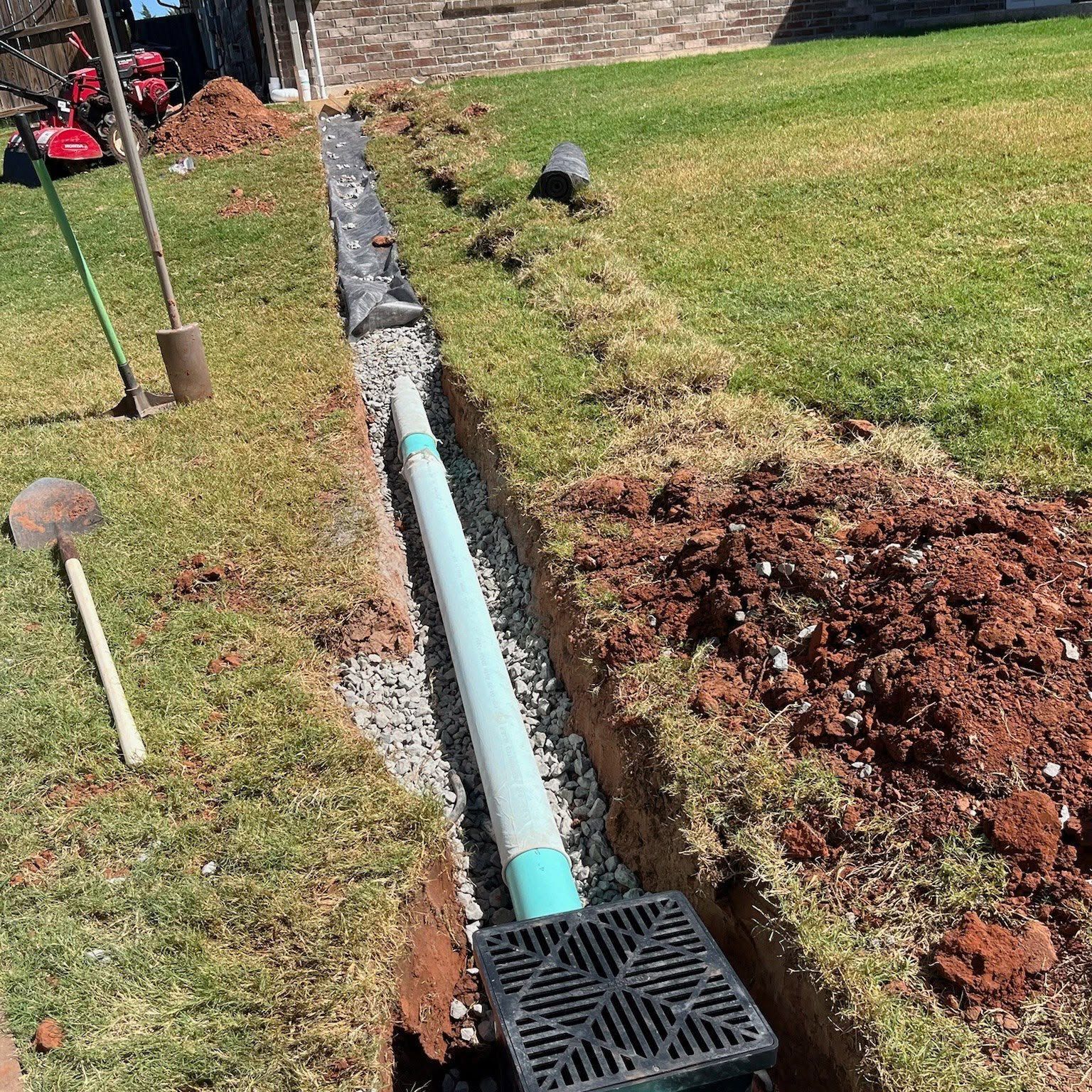 Open trench with blue drainage pipe and black drain grate beside a grassy lawn.